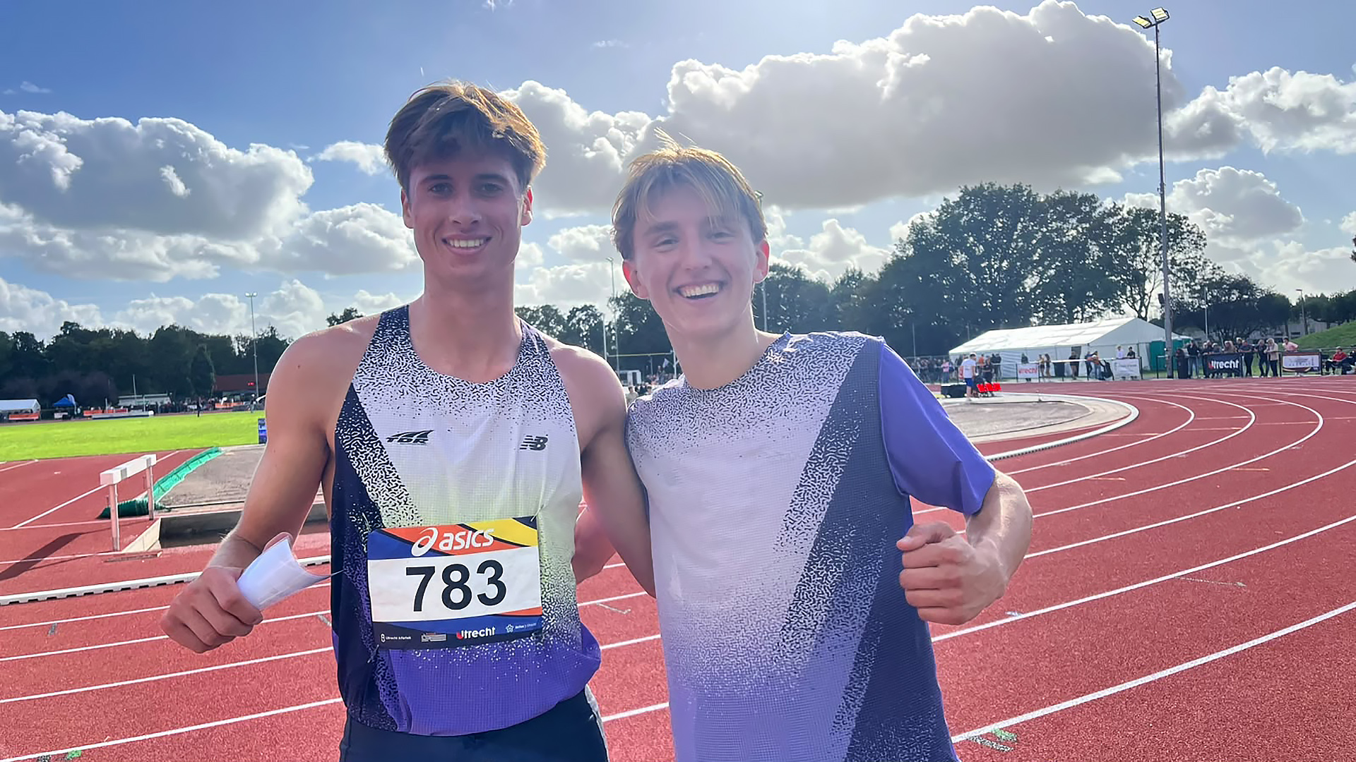 Twee lachende atleten op een atletiekbaan, één met startnummer 783, met bocht van de baan en wolkenlucht op de achtergrond
