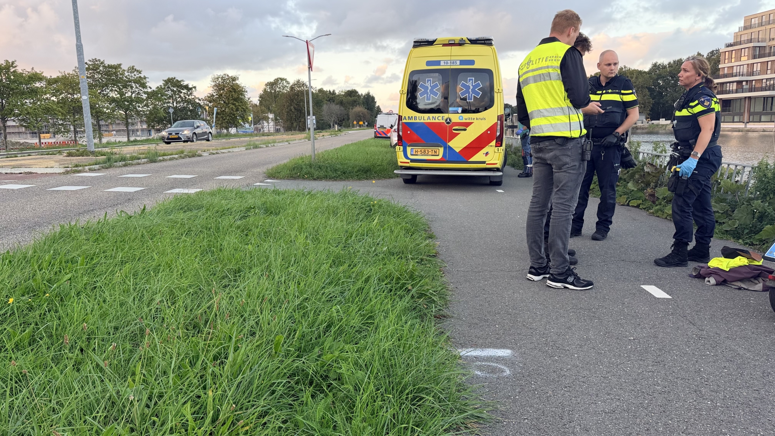 Politieagenten praten naast een geparkeerde ambulance op een fietspad langs het water, met kleding op de grond