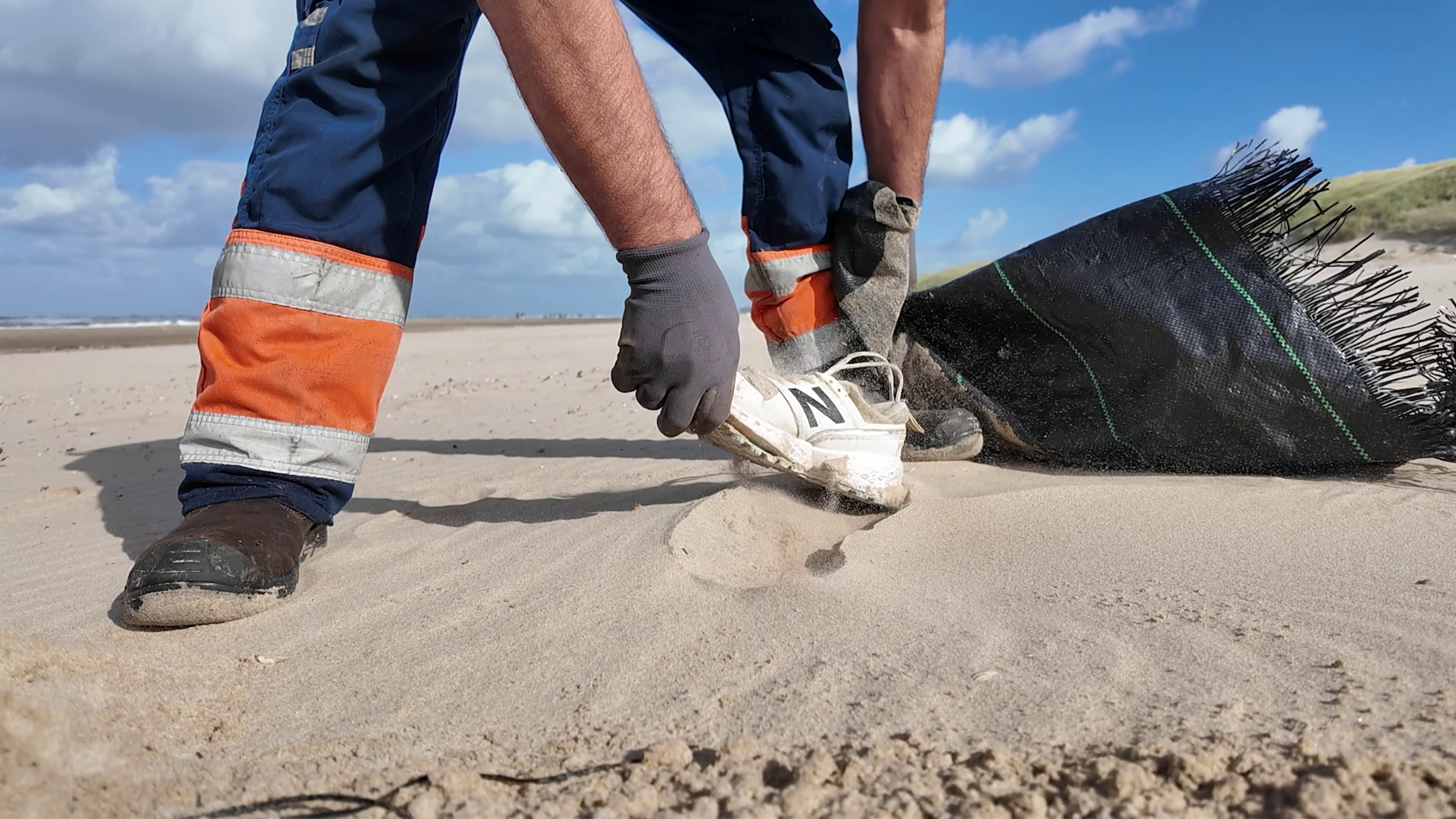 Werker met handschoenen raapt een witte sneaker op van het strand naast een zwarte zandzak
