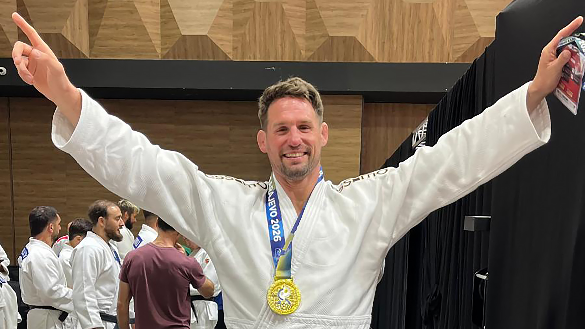 Man in judopak met gouden medaille om zijn nek, armen omhoog in sporthal met andere judoka’s op de achtergrond