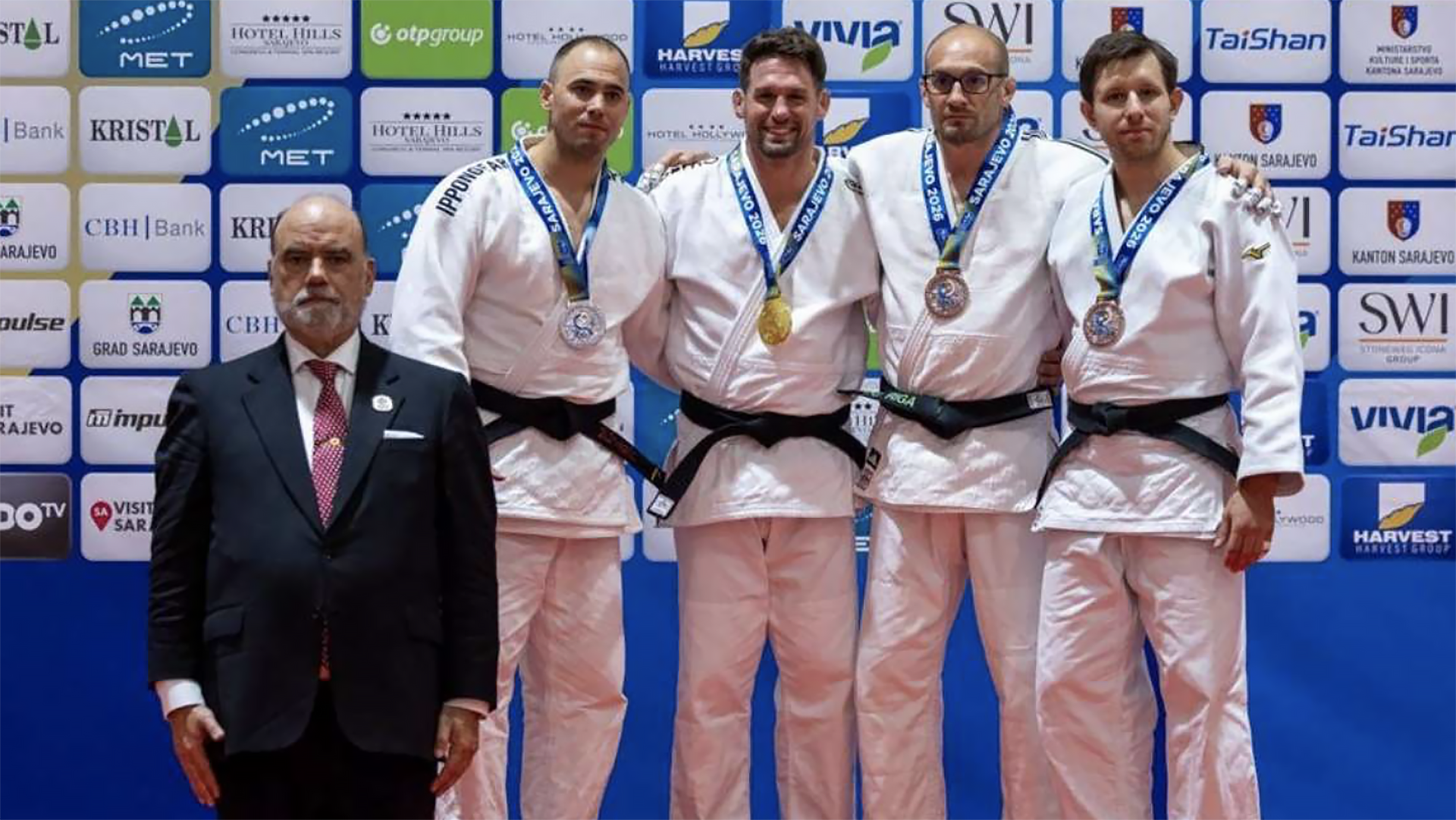 Vier judoka’s in witte gi met medailles poseren op het podium voor een sponsorwand, met een official in pak ervoor