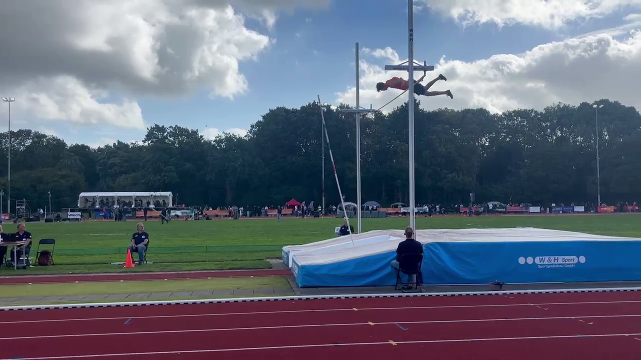 Polsstokhoogspringer zweeft boven de lat op een atletiekbaan, met blauwe landingsmat en toeschouwers op de achtergrond