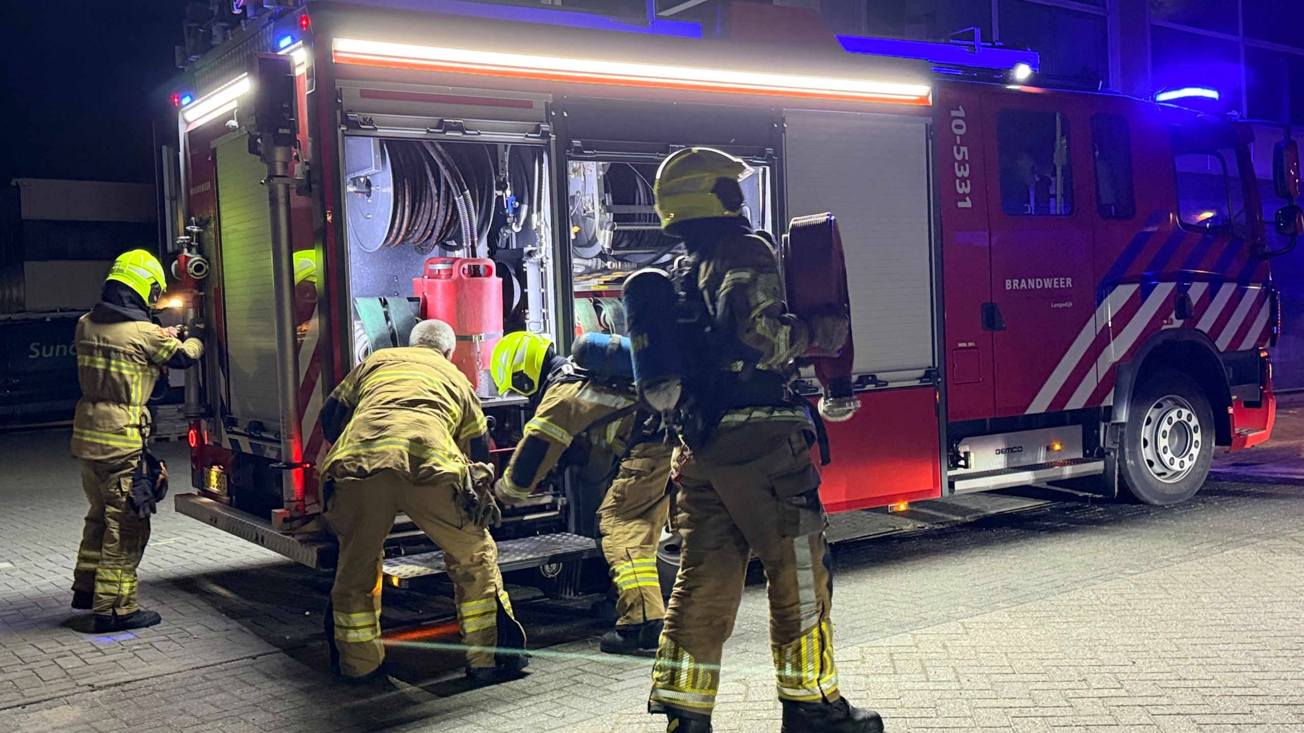 Brandweerlieden bij een brandweerwagen met open compartimenten, terwijl ze slangen en uitrusting pakken in het donker