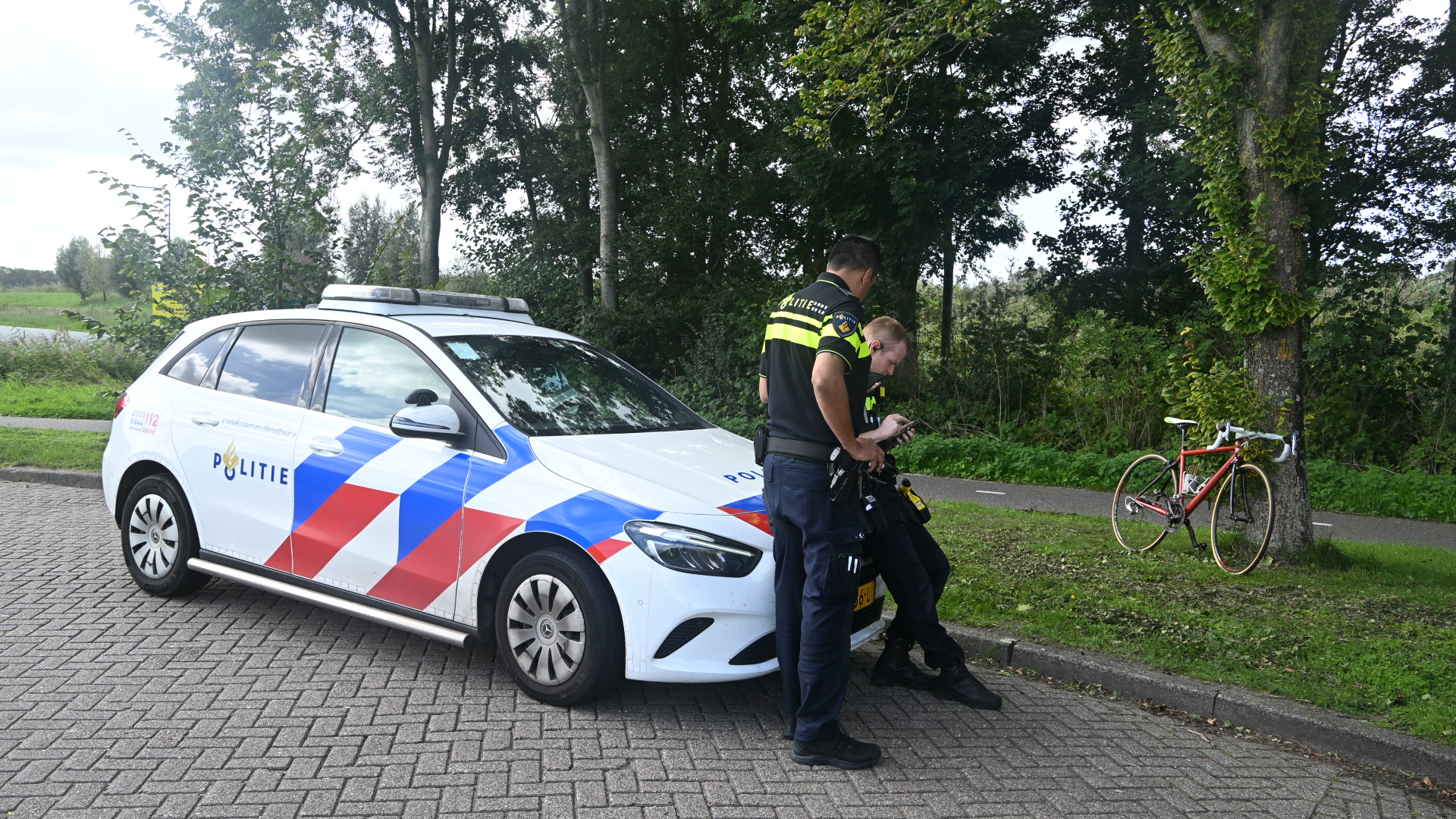 Twee politieagenten staan bij een Nederlandse politieauto en kijken op een telefoon, met een racefiets tegen een boom