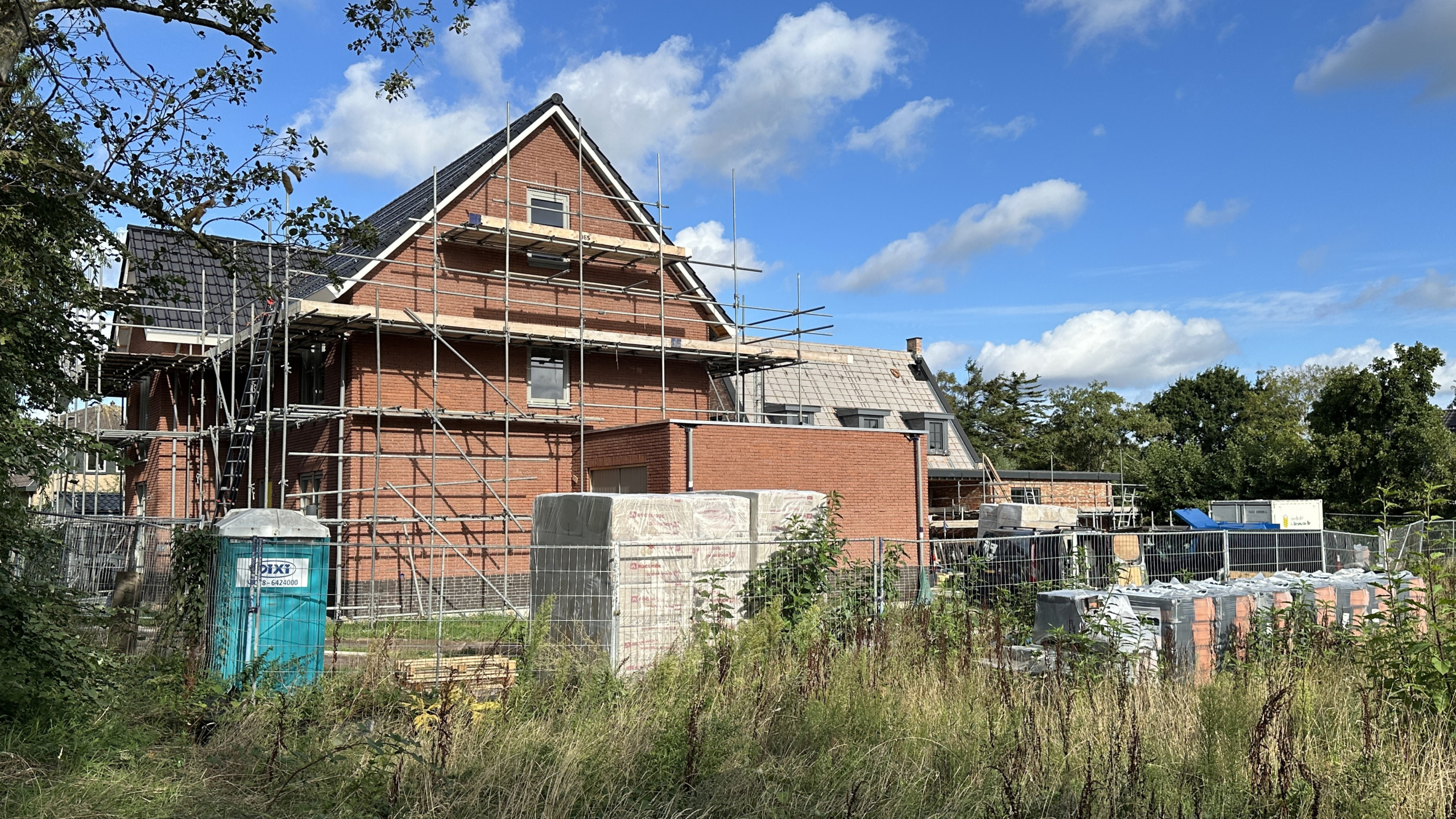 Bakstenen huis in aanbouw met steigers, bouwhekken en stapels bouwmateriaal onder een blauwe lucht