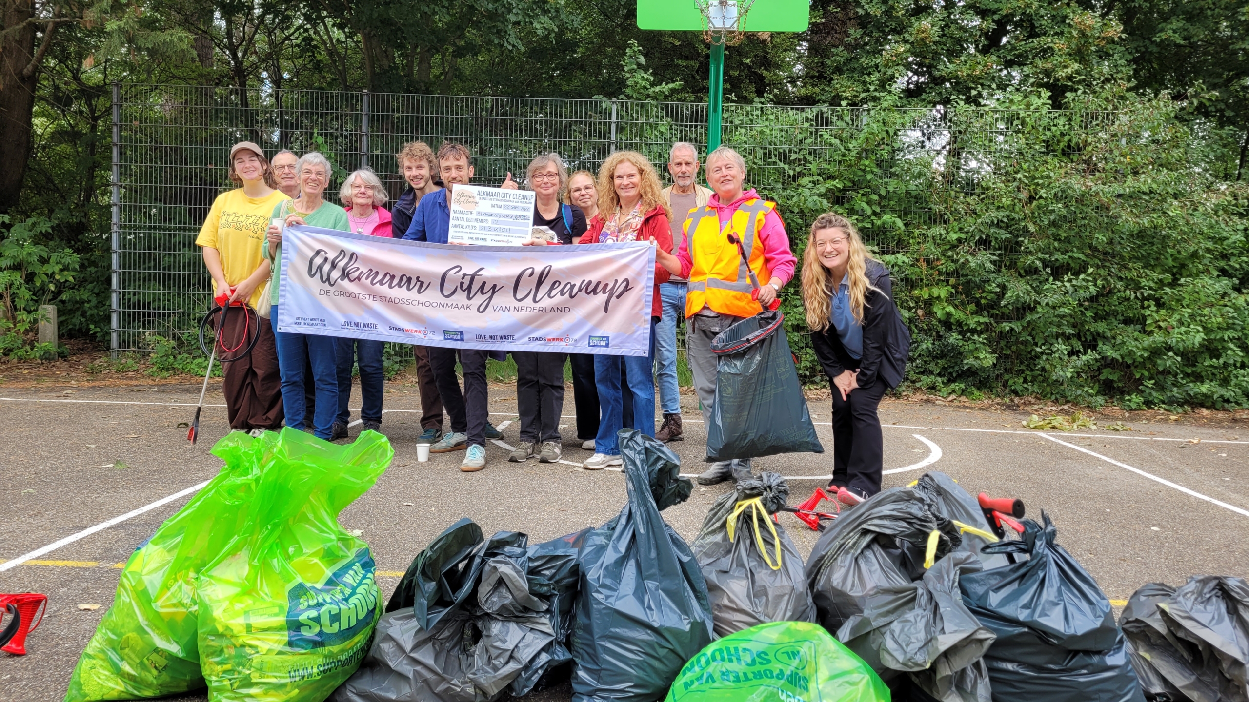 Groep vrijwilligers met spandoek “Alkmaar City Cleanup” poseert op sportveld, met volle vuilniszakken op de voorgrond