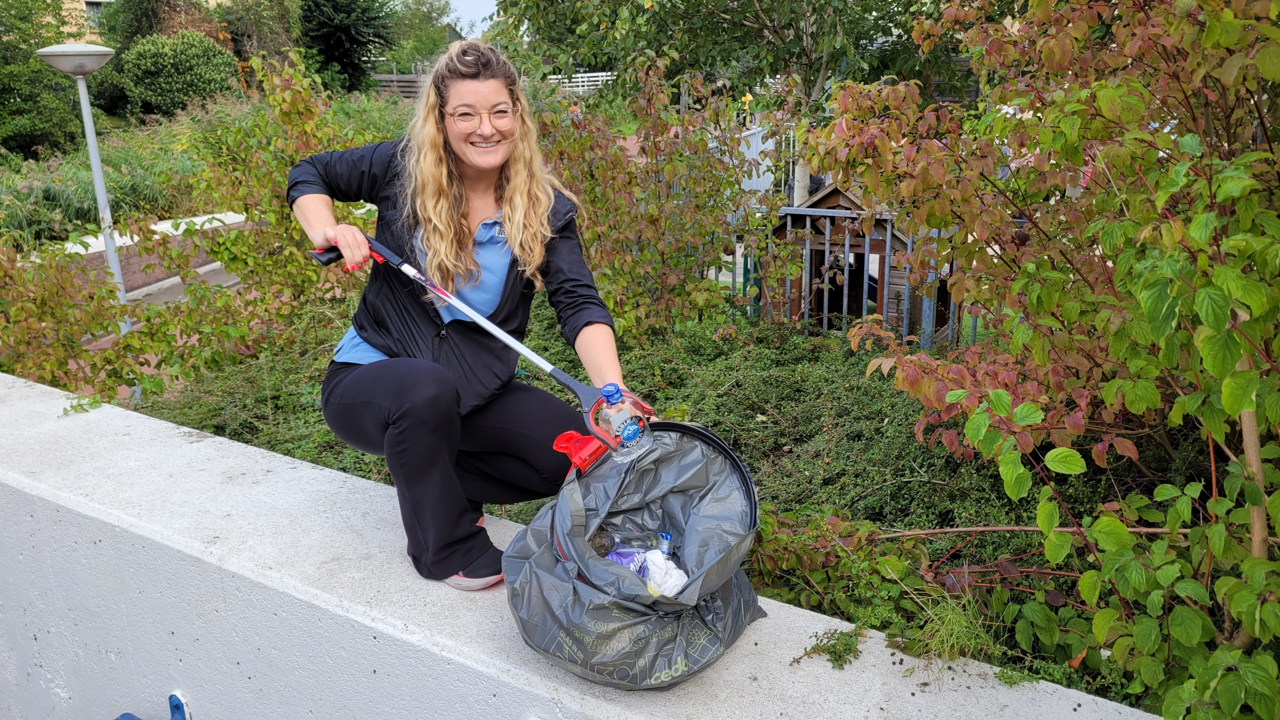 Vrouw in een groen park raapt zwerfafval met een grijper en vult een vuilniszak met plastic flessen