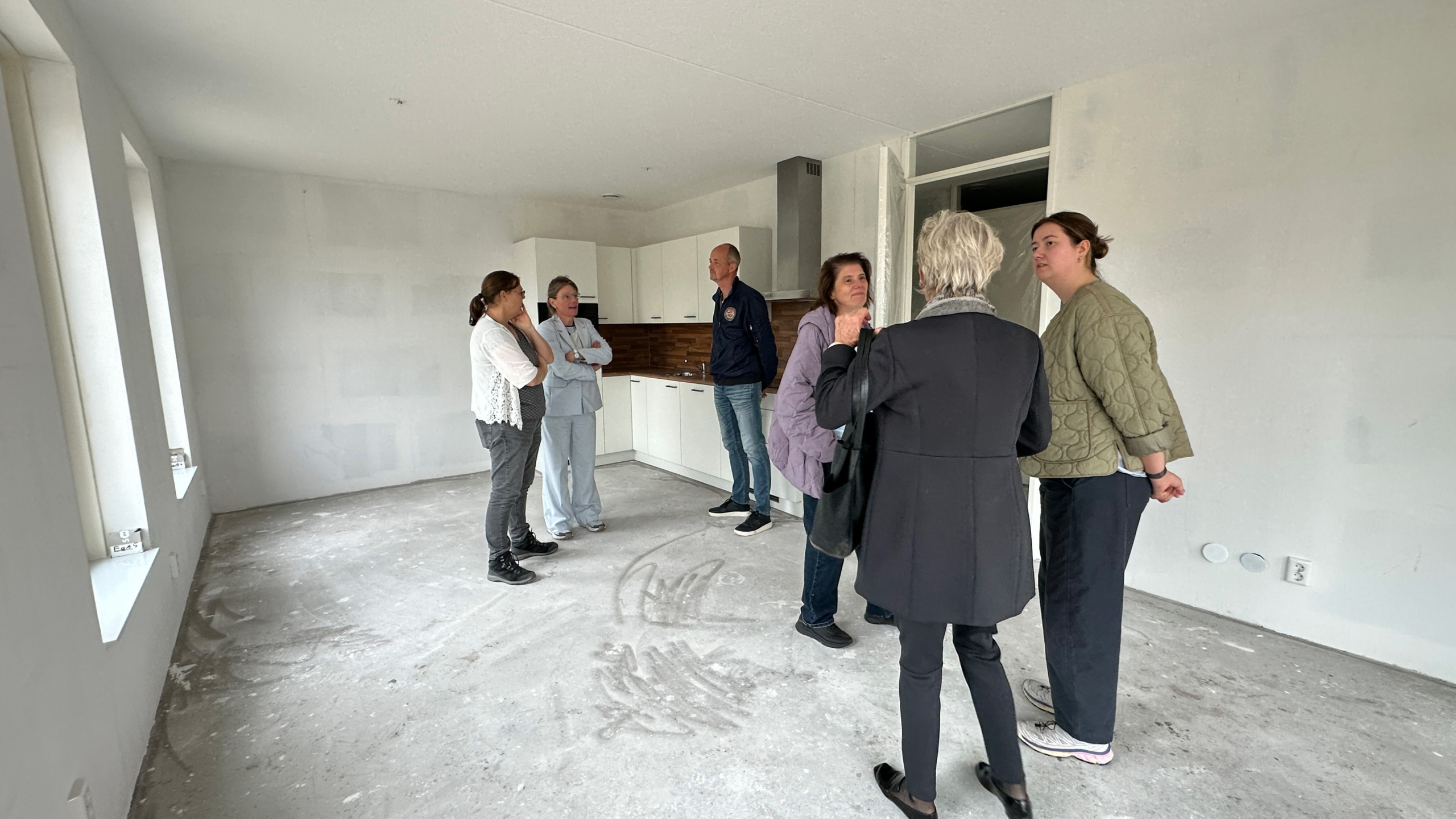Zes mensen overleggen in een lege woning met betonnen vloer en nieuwe witte keuken in de hoek