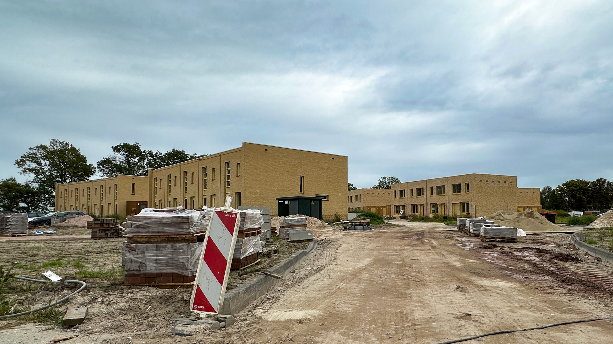 Bouwplaats met nieuwe bakstenen gebouwen, pallets met stenen en een rood-wit wegafzetbord langs een modderige weg