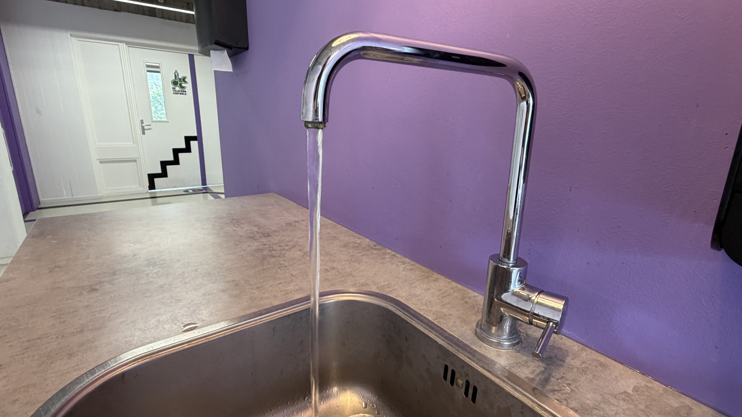 Chromen keukenkraan met stromend water boven een roestvrijstalen gootsteen, tegen een paarse muur
