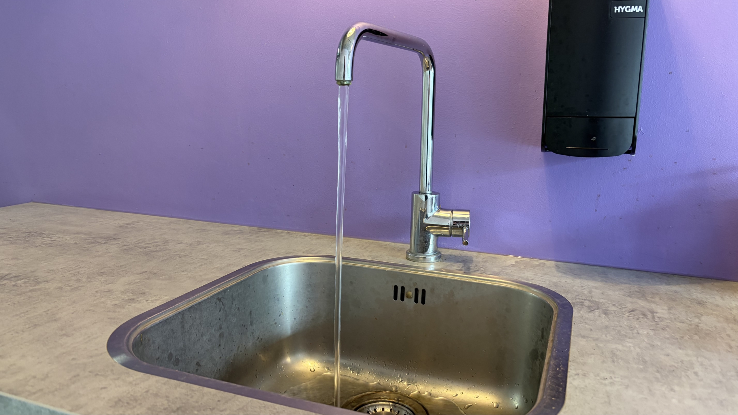 Waterstraal uit een chromen keukenkraan in een roestvrijstalen spoelbak, met paarse muur op de achtergrond