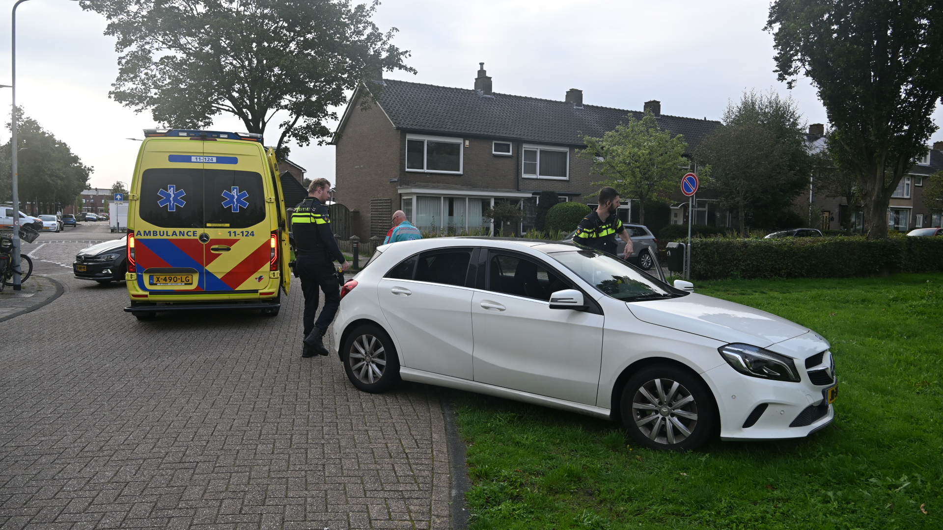 Ambulance en twee agenten bij witte auto die deels op het gras staat in een woonwijk