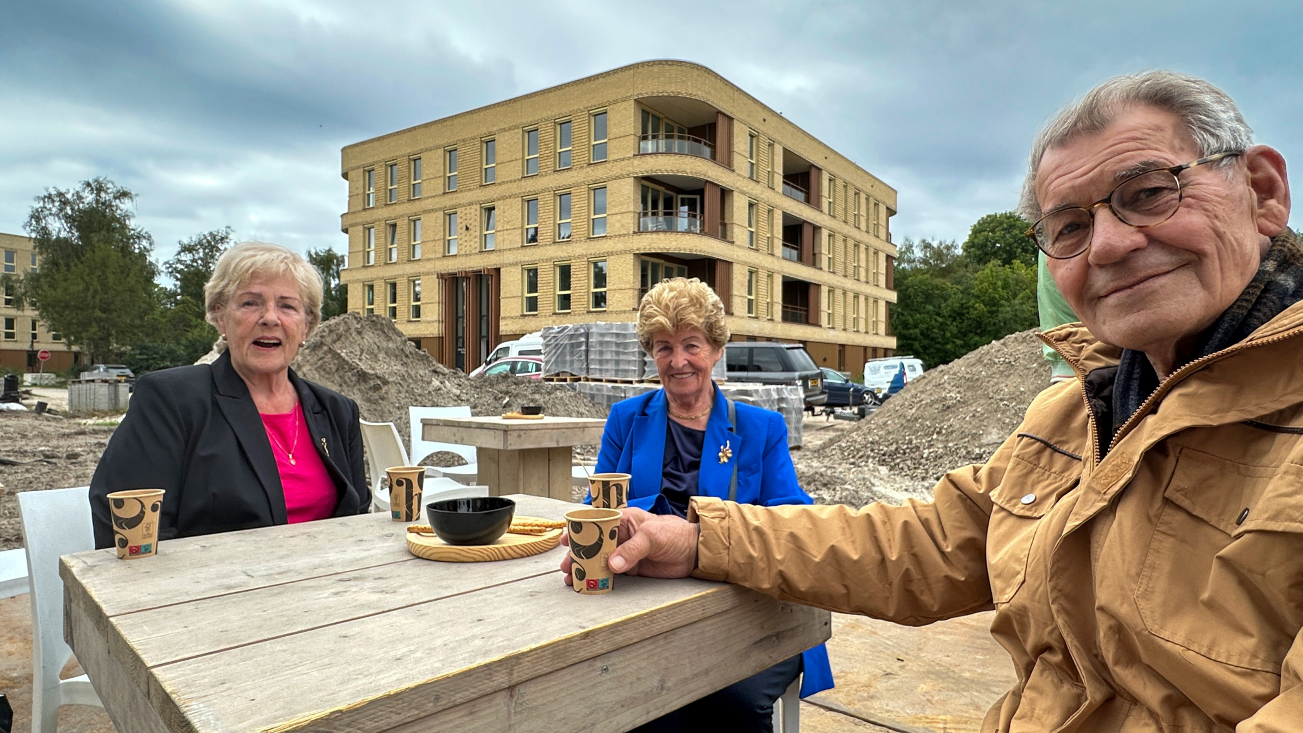 Drie ouderen drinken koffie aan een houten tafel bij een bouwplaats, met nieuw appartementencomplex op de achtergrond