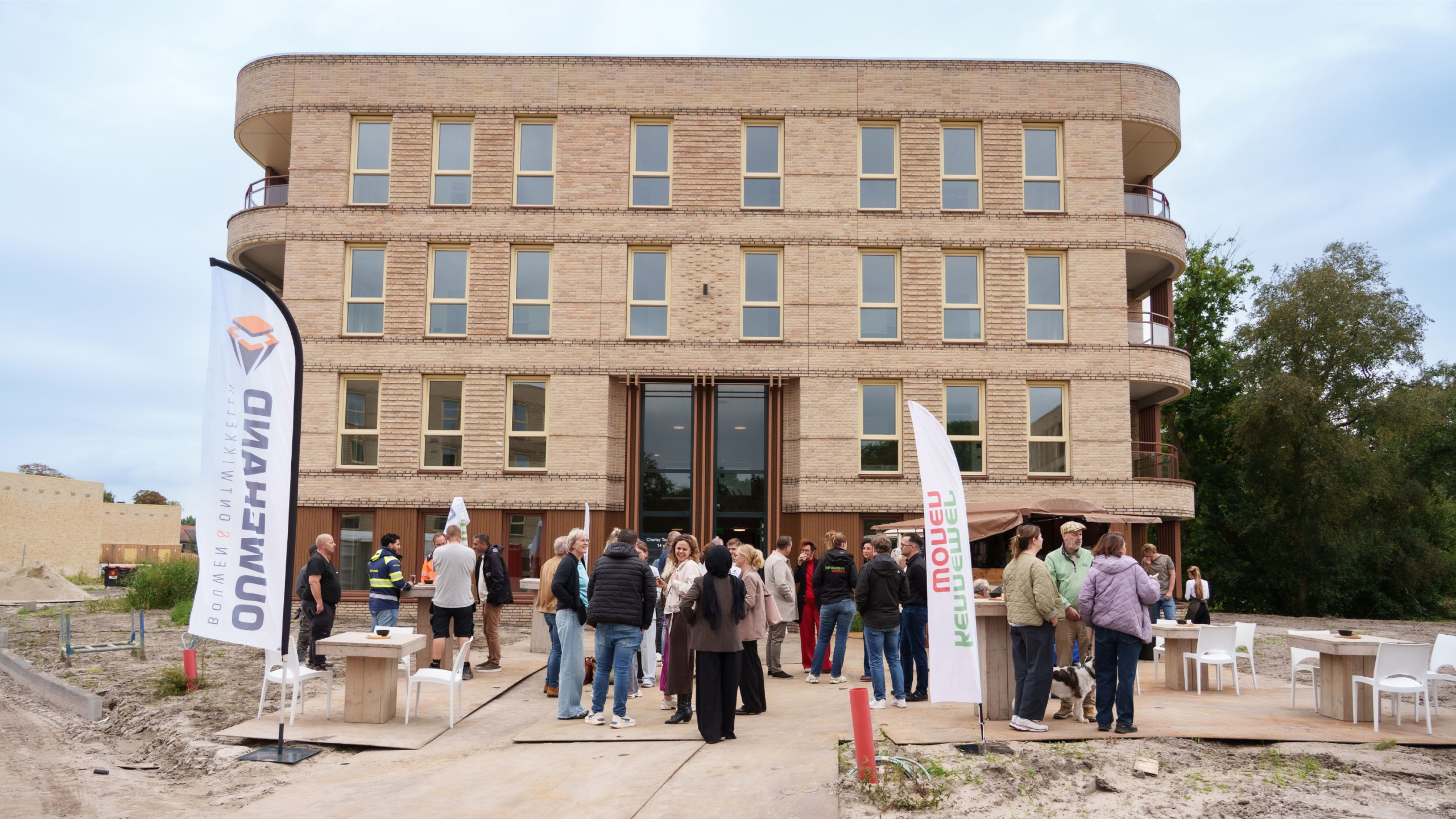 Groep mensen bij opening van nieuw bakstenen gebouw, met vlaggen “Oomen” en “reule deuel” voor de ingang
