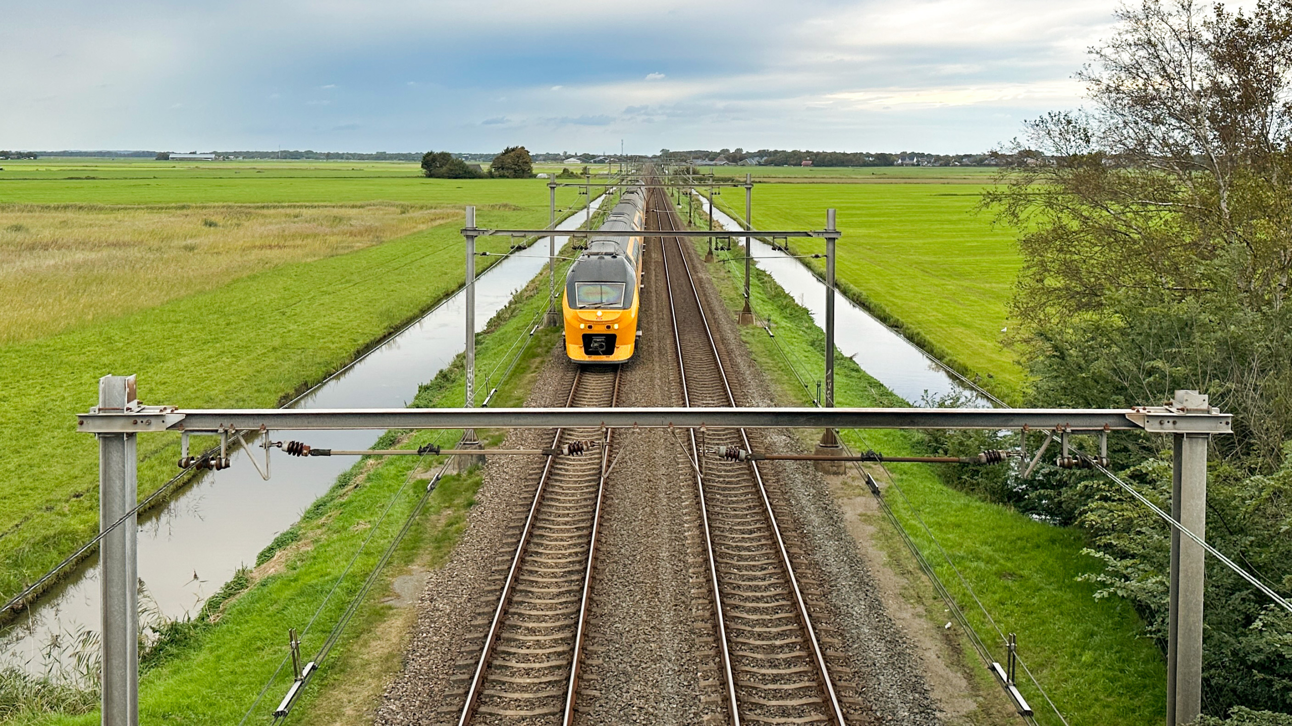 Gele trein rijdt over dubbel spoor door groen polderlandschap, met sloten langs het spoor, gezien vanaf een brug