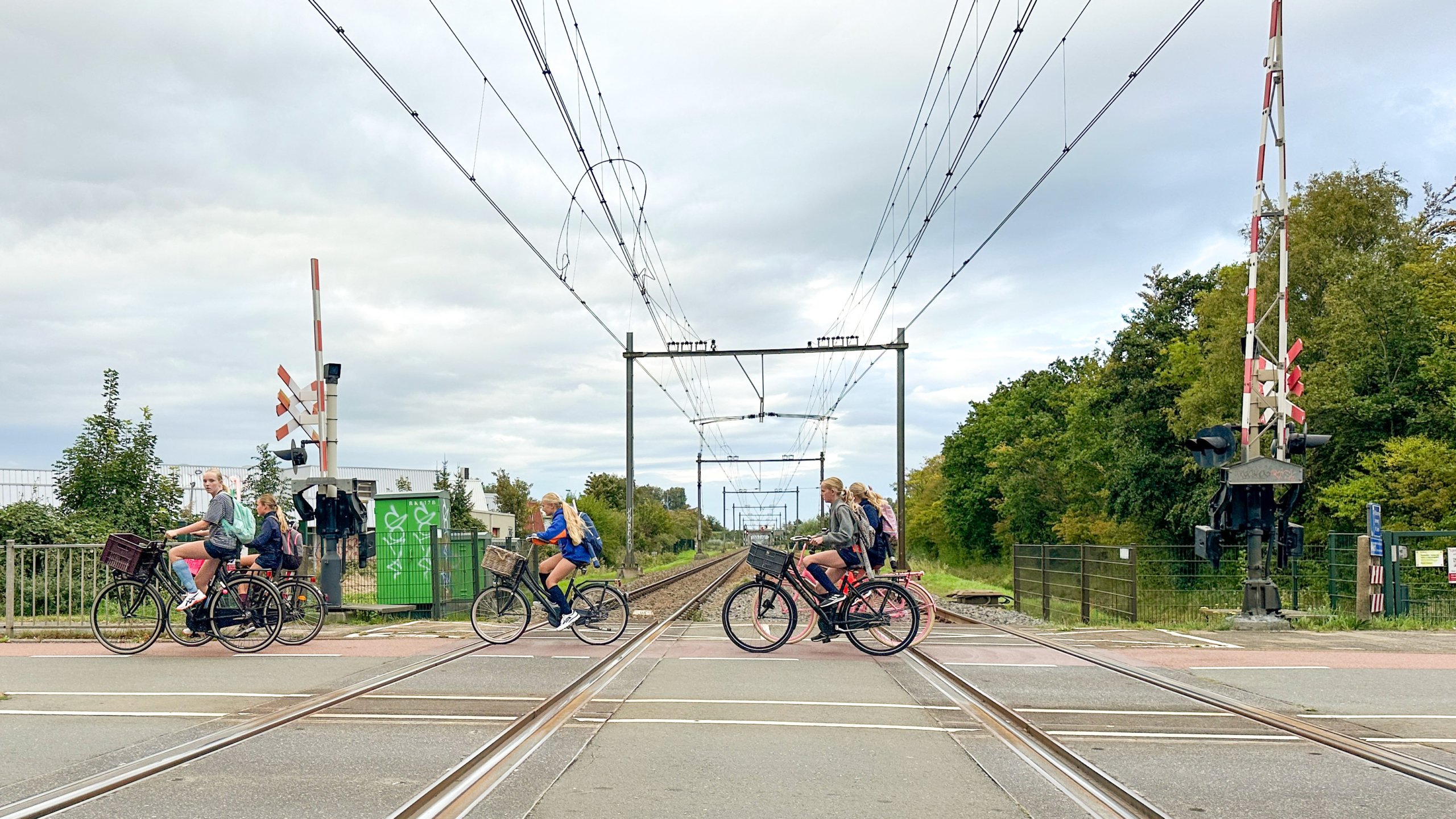 Fietsers steken een spoorwegovergang over met bovenleidingen en bomen langs het spoor onder een bewolkte lucht