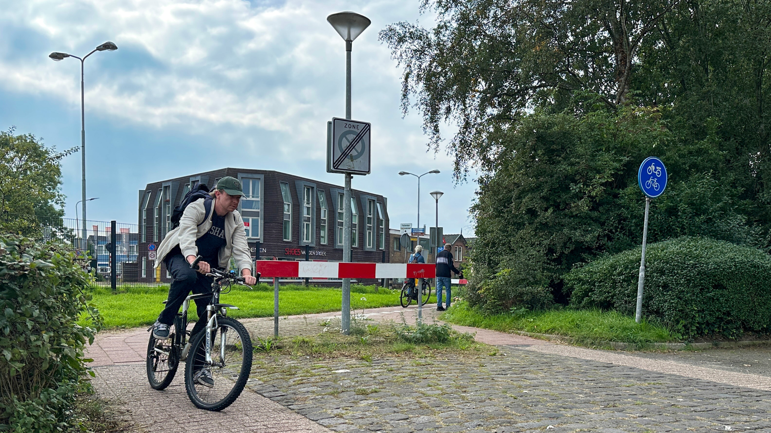 Man fietst met rugzak over fietspad langs rood-witte slagboom, met verkeersborden en gebouw op de achtergrond