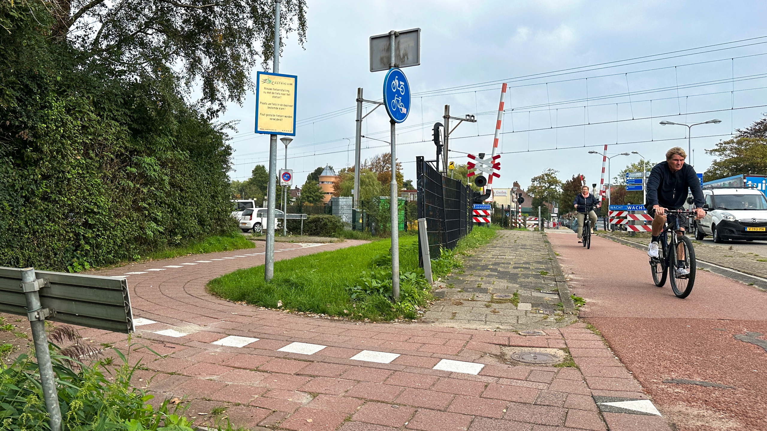 Fietspad langs spoorwegovergang met slagbomen en waarschuwingsborden, met twee fietsers en wachtende auto’s