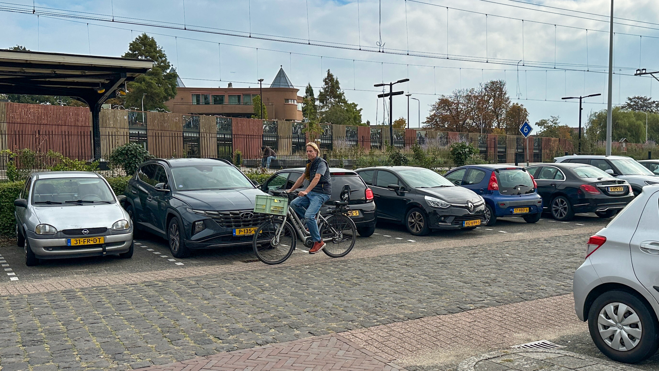 Man op fiets met groene krat rijdt langs geparkeerde auto’s bij een station, met bovenleiding en hek op de achtergrond