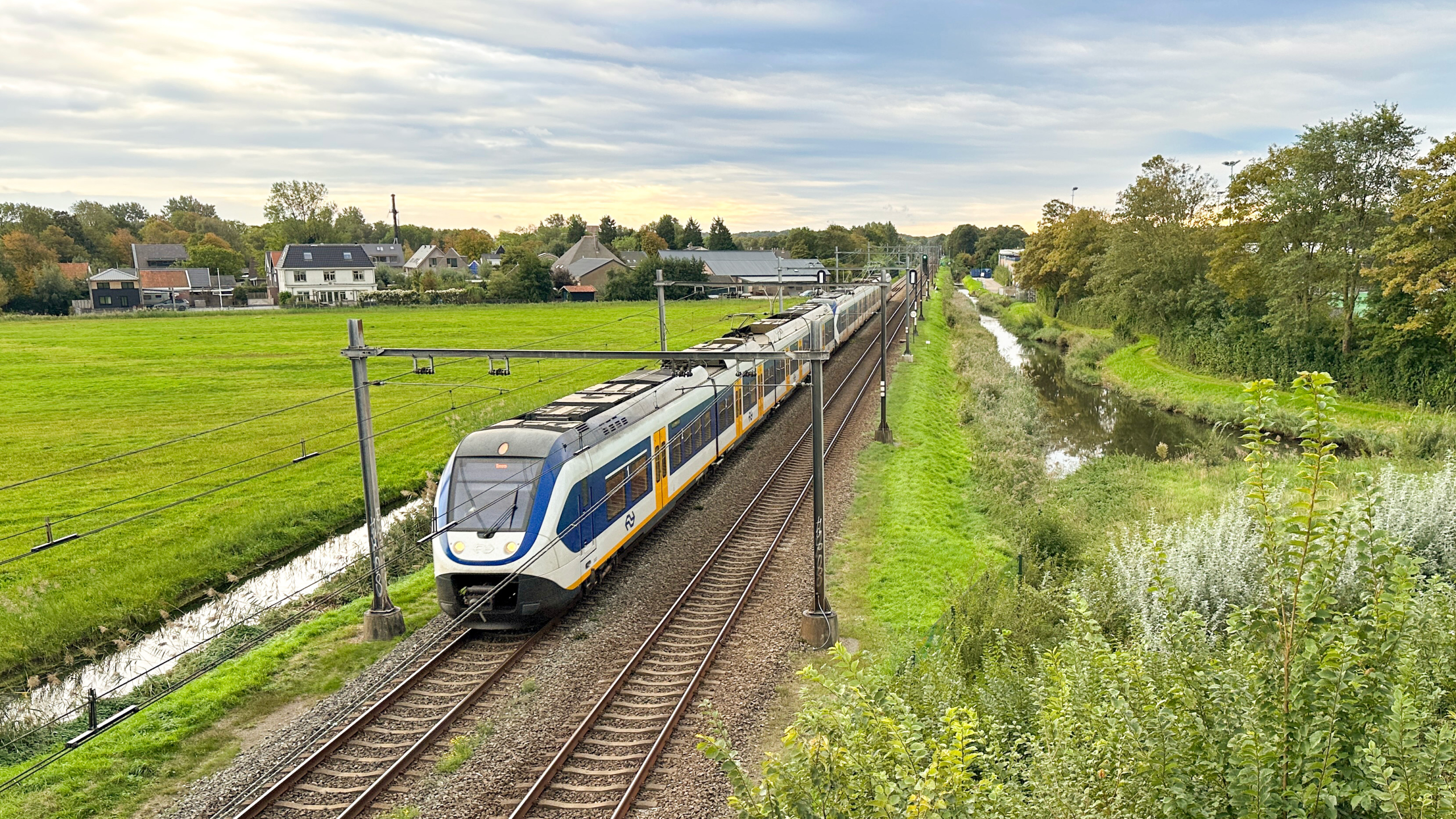 Dubbeldekker trein rijdt over spoor langs groen weiland en kanaal, met huizen en bomen op de achtergrond