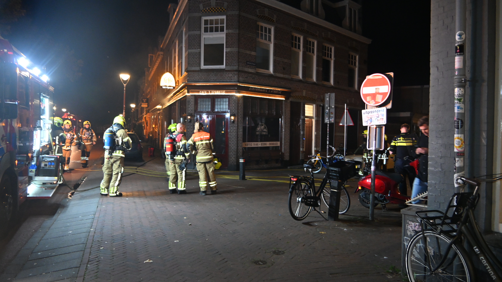 Brandweerlieden en politie op straat bij een hoekpand in de nacht, met brandweerwagen, slangen en fietsen op de voorgrond