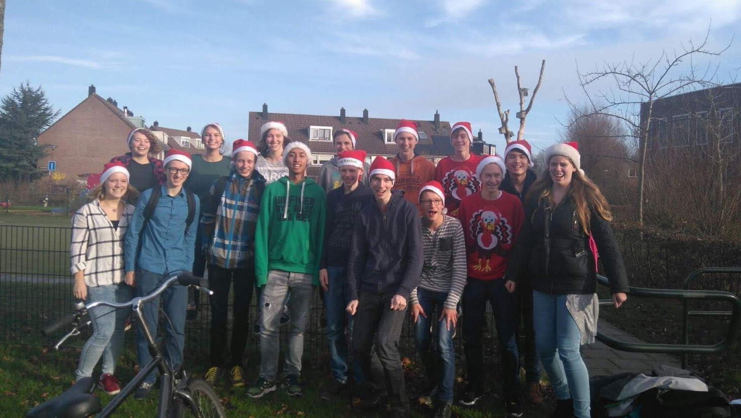 Groep jongeren met kerstmutsen poseert lachend buiten in een woonwijk, met een fiets op de voorgrond