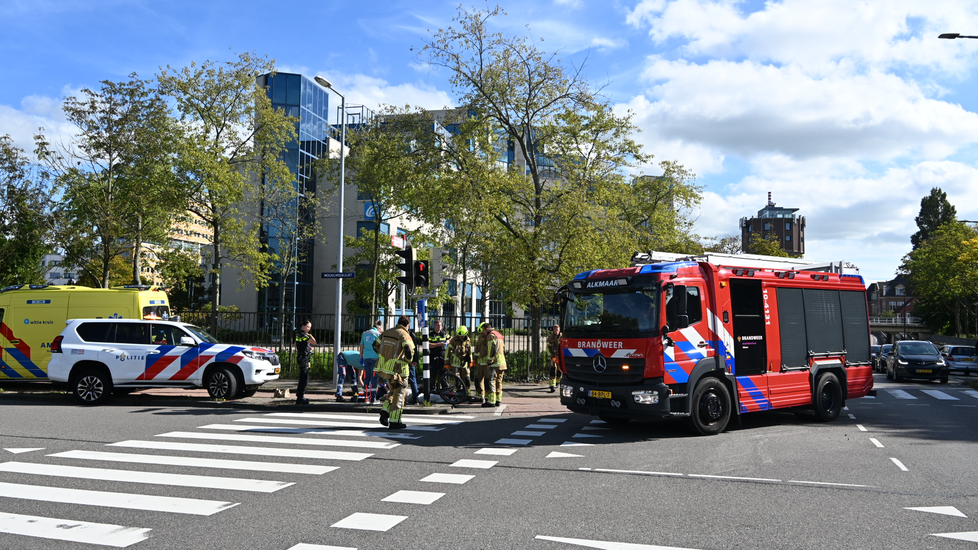 Brandweerwagen, politieauto en ambulance staan bij een kruispunt terwijl hulpverleners iemand op straat verzorgen