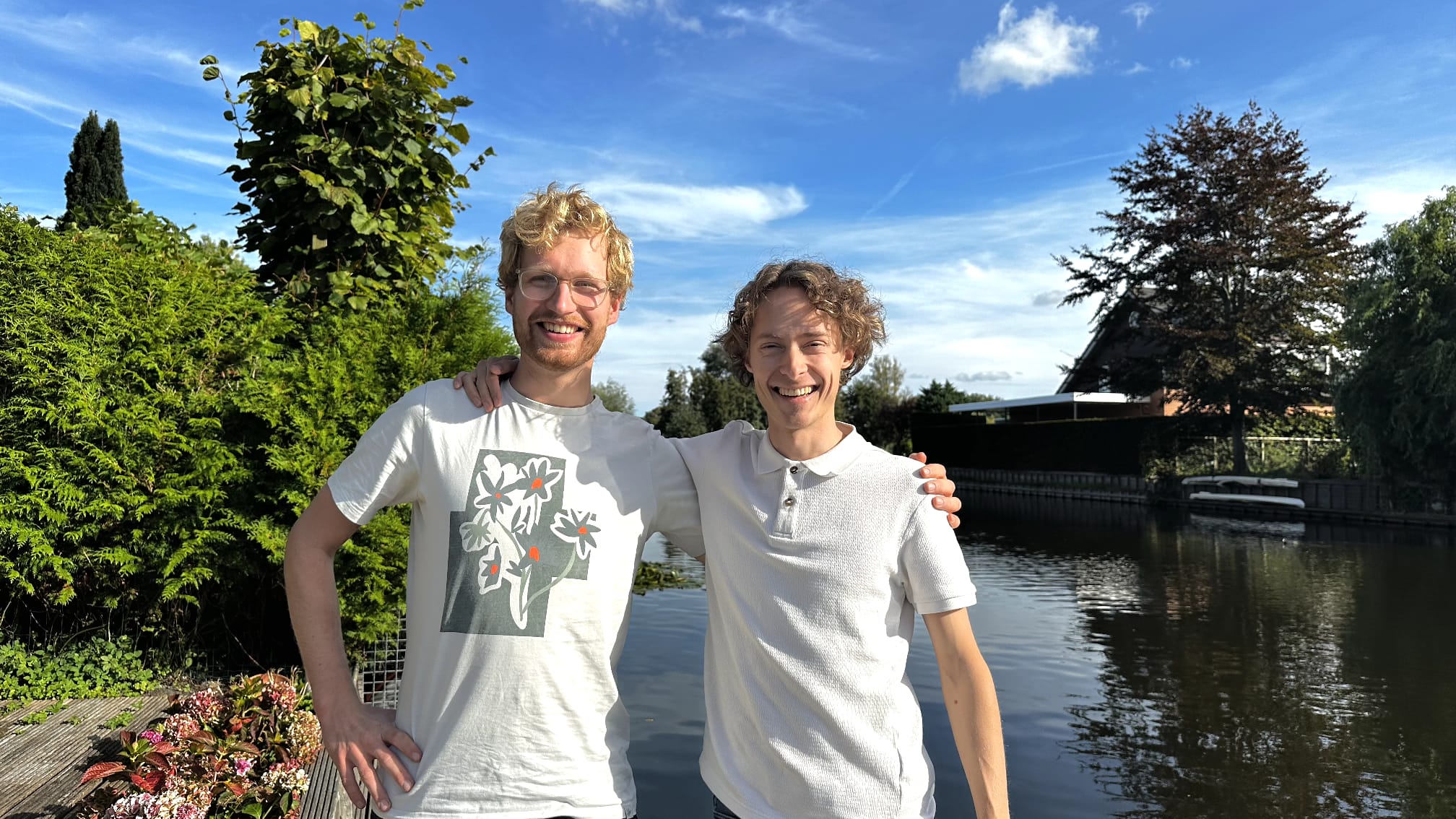 Twee lachende mannen met hun armen om elkaar, staand bij een vijver met bomen en blauwe lucht op de achtergrond
