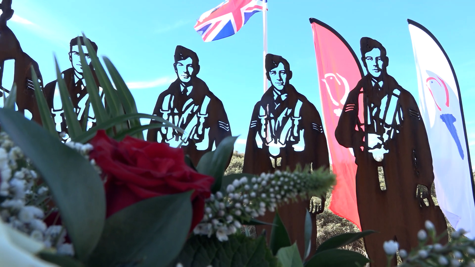 Stalen silhouetten van piloten met parachuteharnas bij een herdenking, met bloemen op de voorgrond en vlaggen erachter
