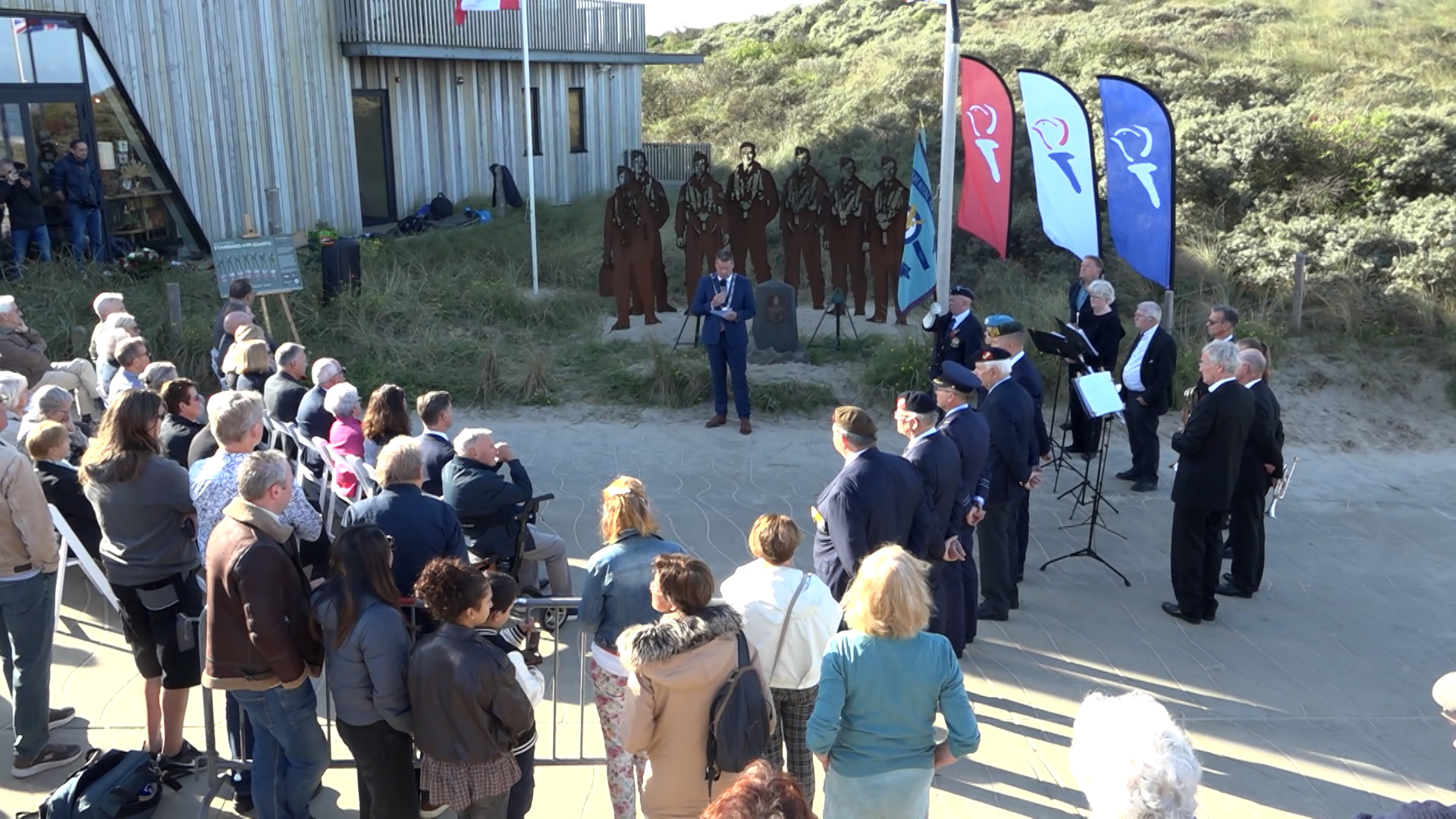 Bijeenkomst bij een monument in de duinen, met spreker, publiek en fanfare naast vlaggen