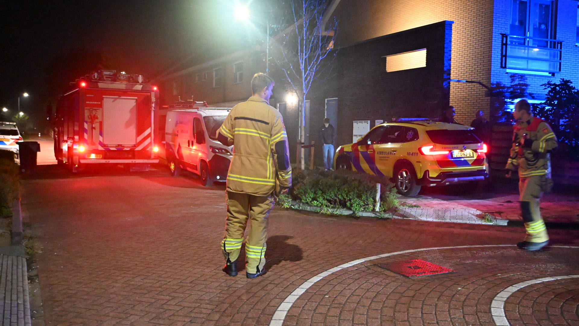 Brandweer en ambulance met zwaailichten bij een woongebouw ’s nachts, met brandweerlieden op straat