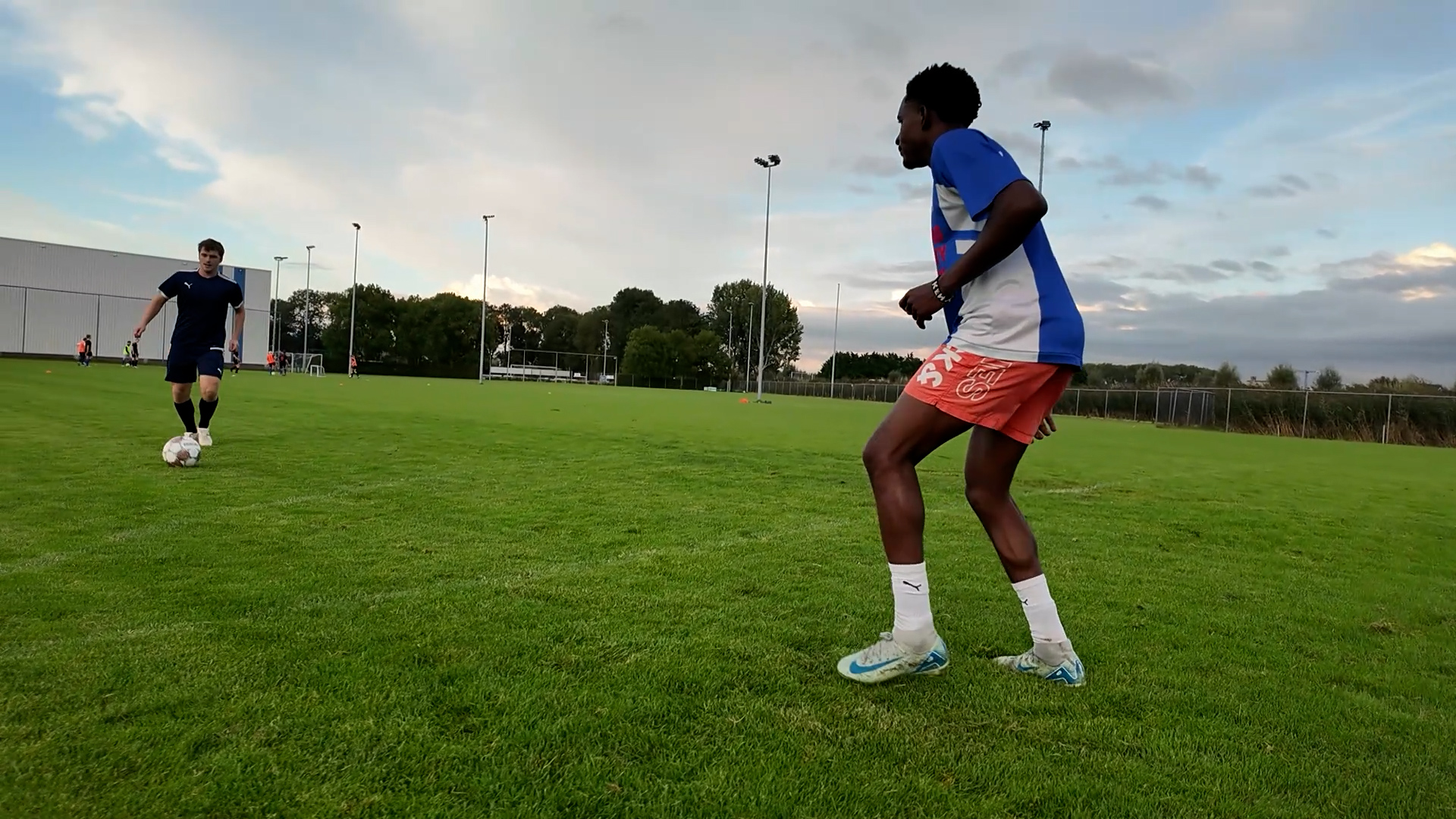Twee voetballers op een grasveld tijdens training, één met bal en de ander klaar om te verdedigen