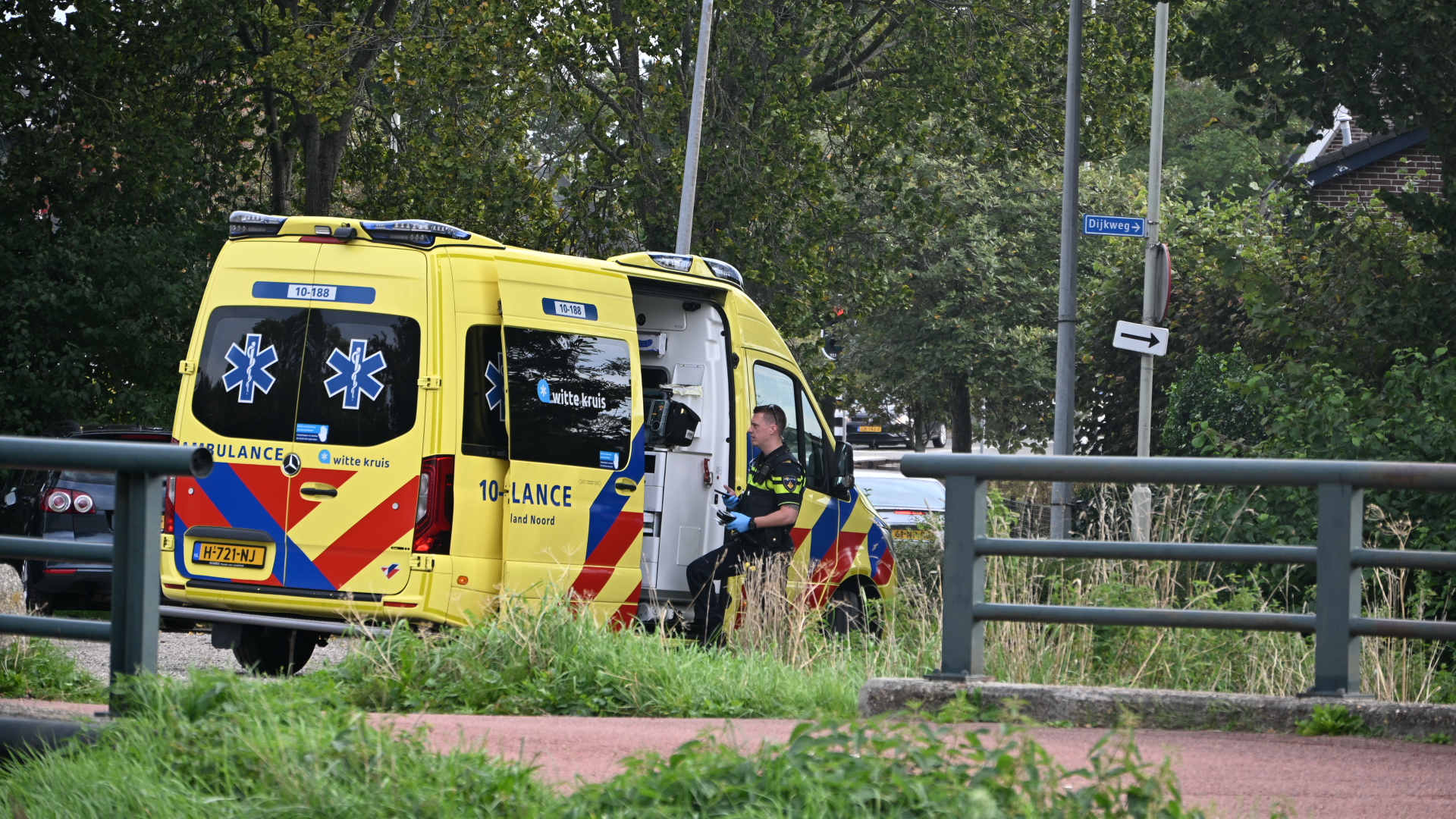 Gele ambulance met open zijdeur; politieagent staat ernaast langs een weg met groen en bordje Dijkweg