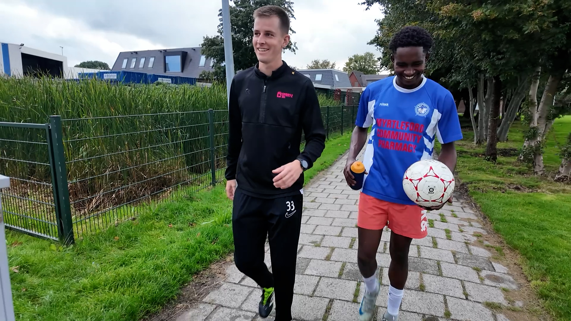Twee mannen lopen lachend over een stoep; één in trainingspak, de ander in voetbalshirt met bal en bidon