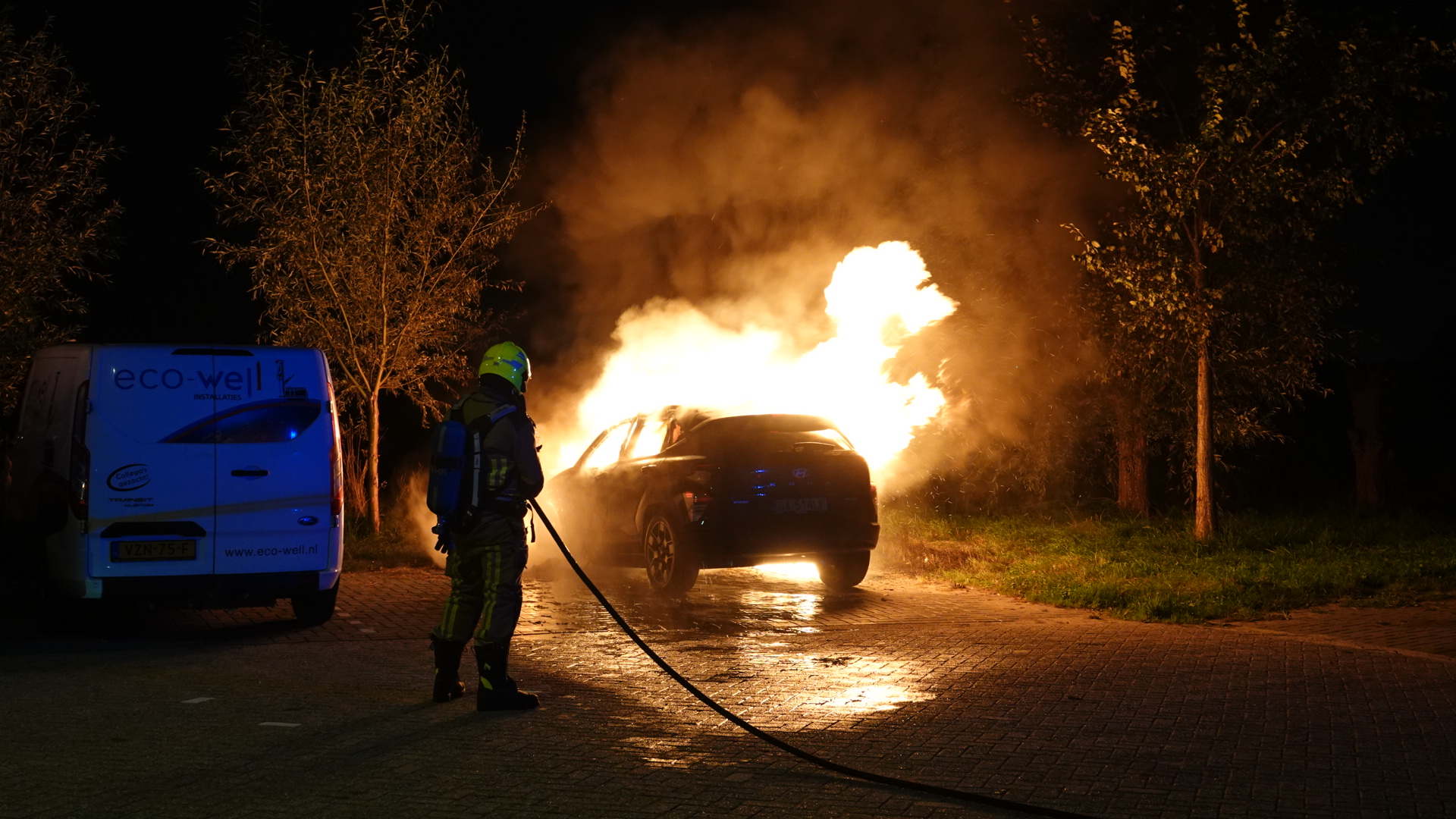Brandweerman blust een brandende auto met grote vlammen en rook op een donkere weg naast een bestelbus