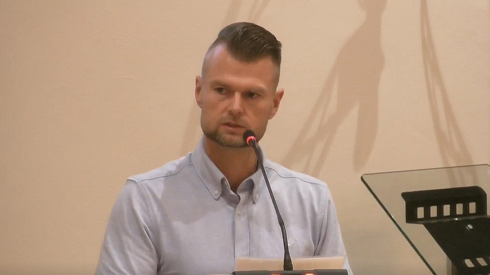 Man met opgeschoren kapsel spreekt in een microfoon tijdens een bijeenkomst, met glasplaat van lessenaar rechts
