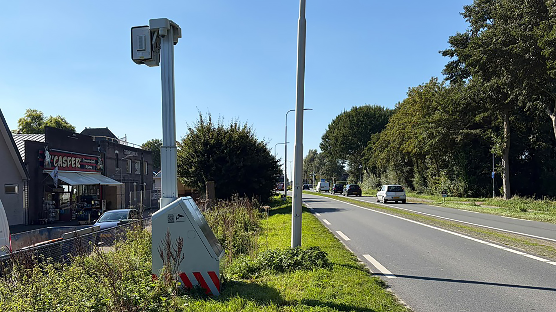 Flitspaal langs een provinciale weg met passerende auto’s, naast groen bermgebied en gebouwen met het bord Casper