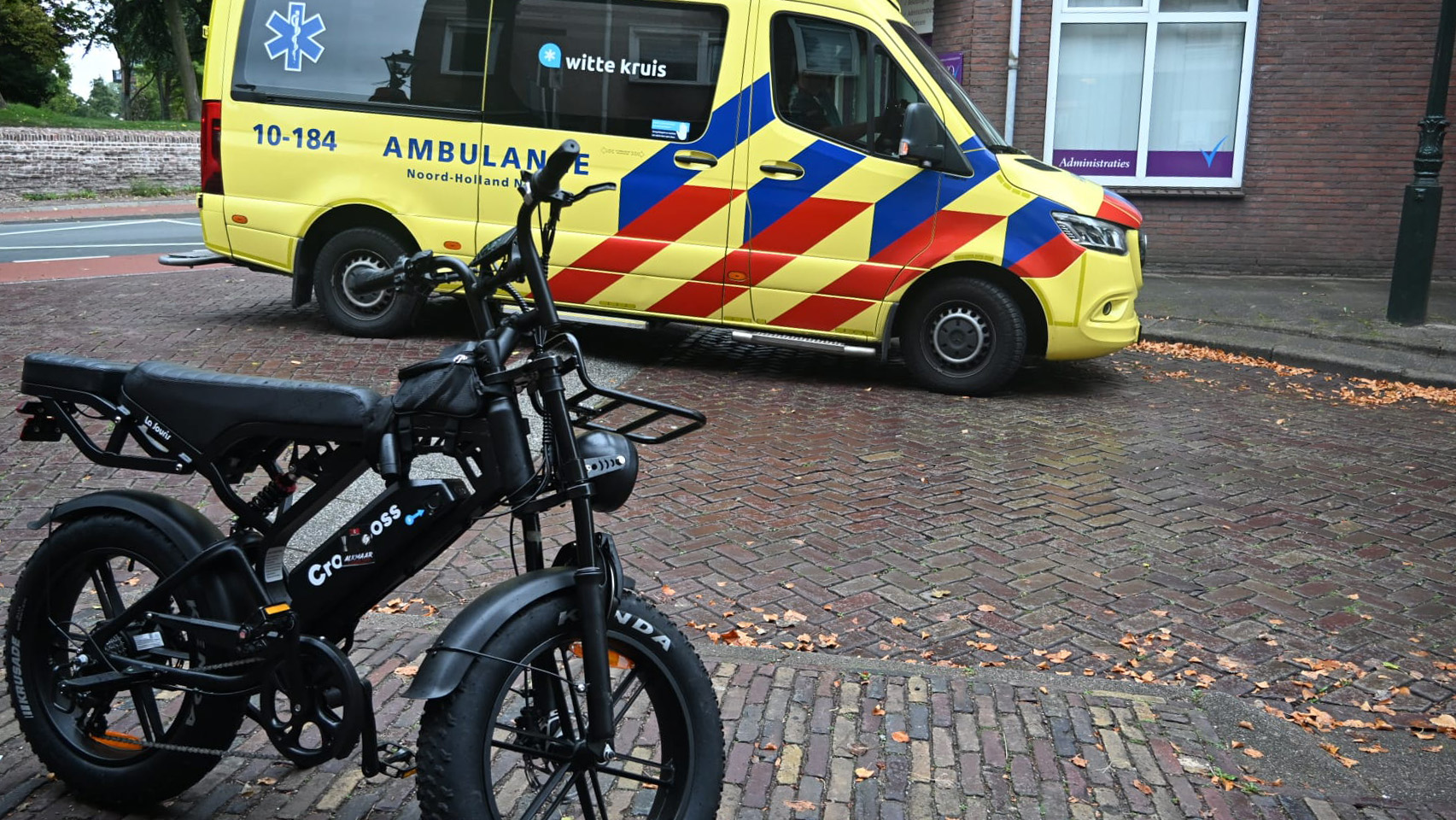 Zwarte elektrische fiets op bakstenen straat, met gele ambulance van Witte Kruis op de achtergrond
