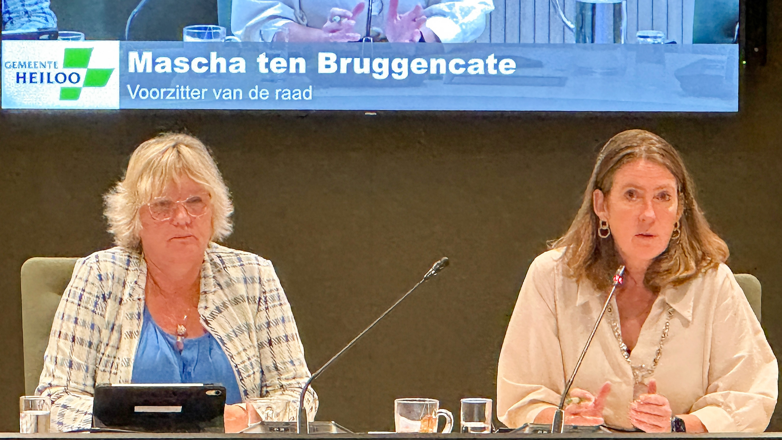 Twee vrouwen aan tafel tijdens raadsvergadering; naambord: Mascha ten Bruggencate, voorzitter van de raad (Gemeente Heiloo)