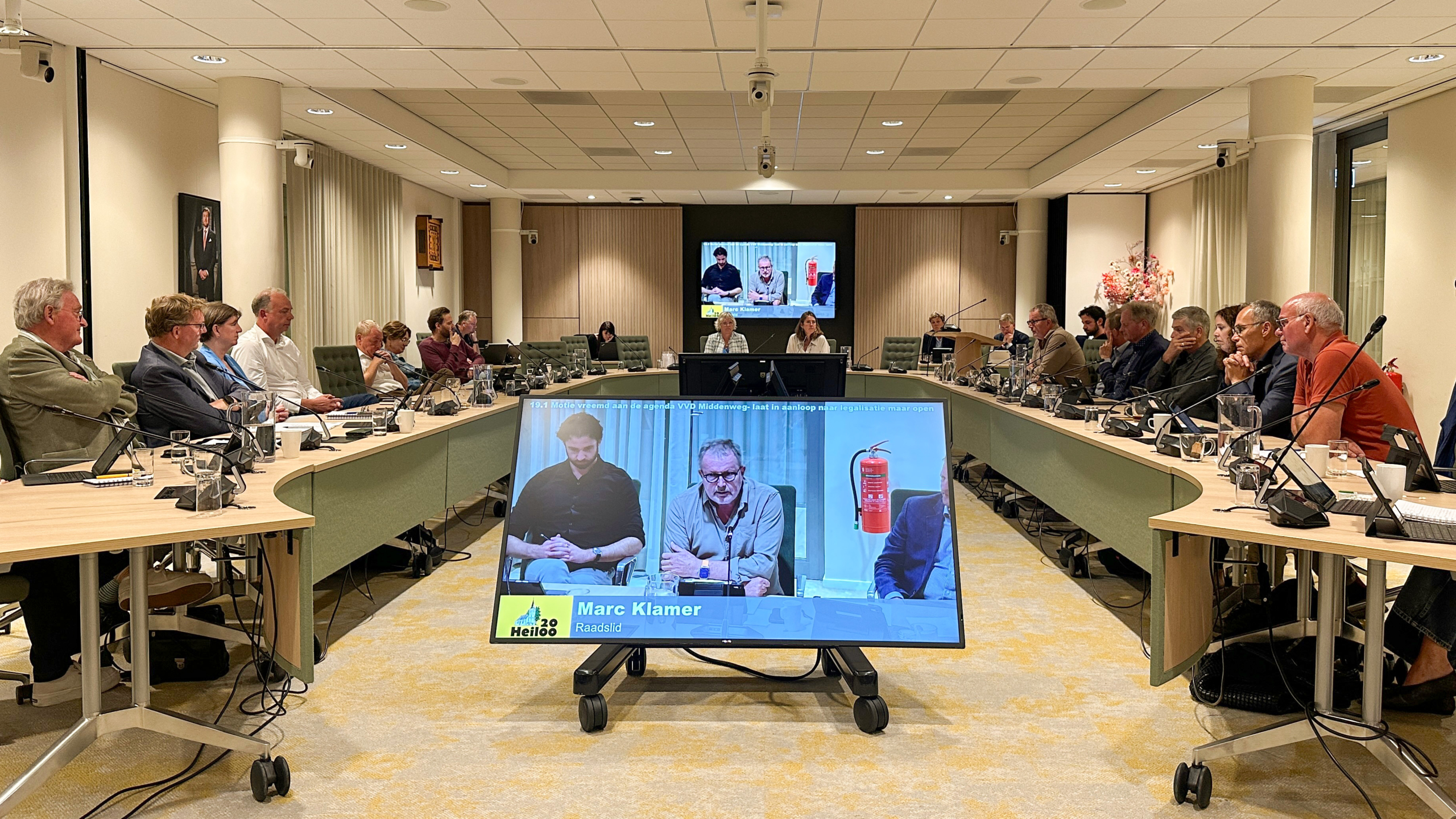 Raadsvergadering in vergaderzaal met raadsleden aan U‑vormige tafels en een scherm met live videobijdrage