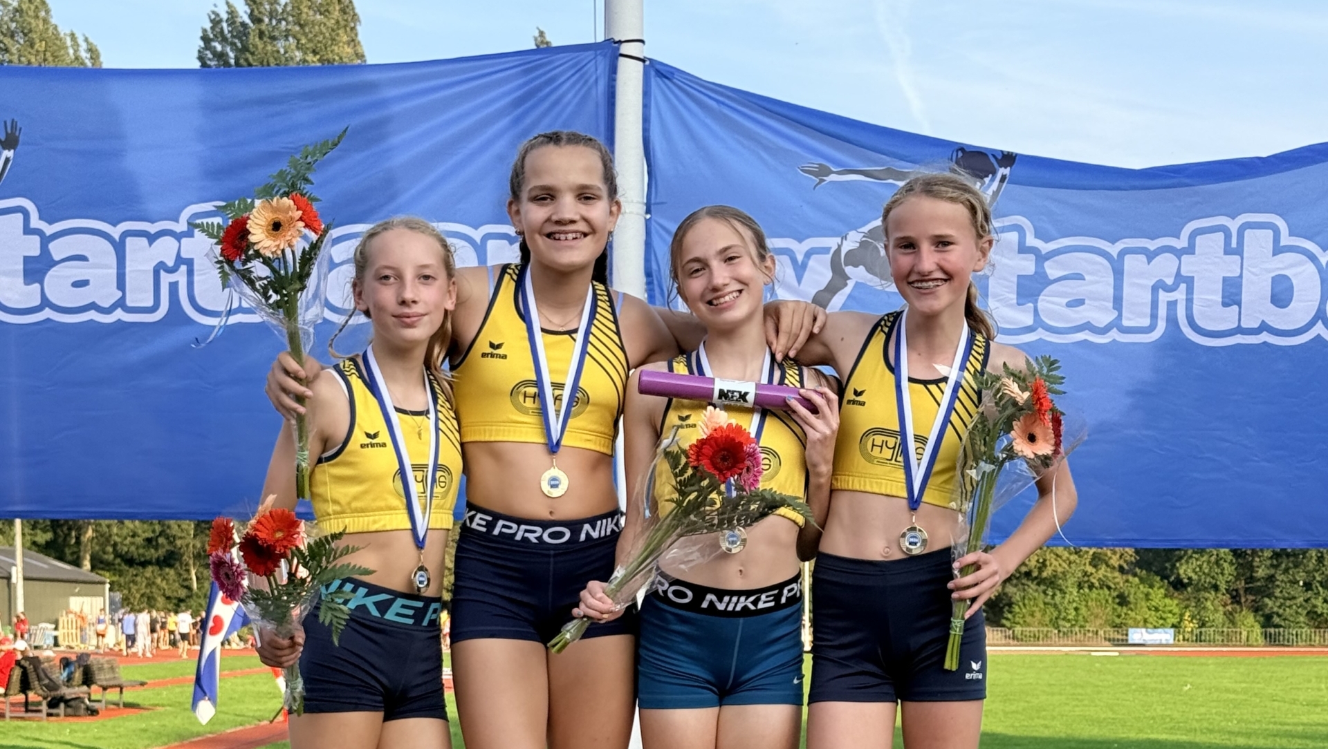 Vier jonge atletes op het podium met medailles, bloemen en estafettestokje bij een atletiekbaan