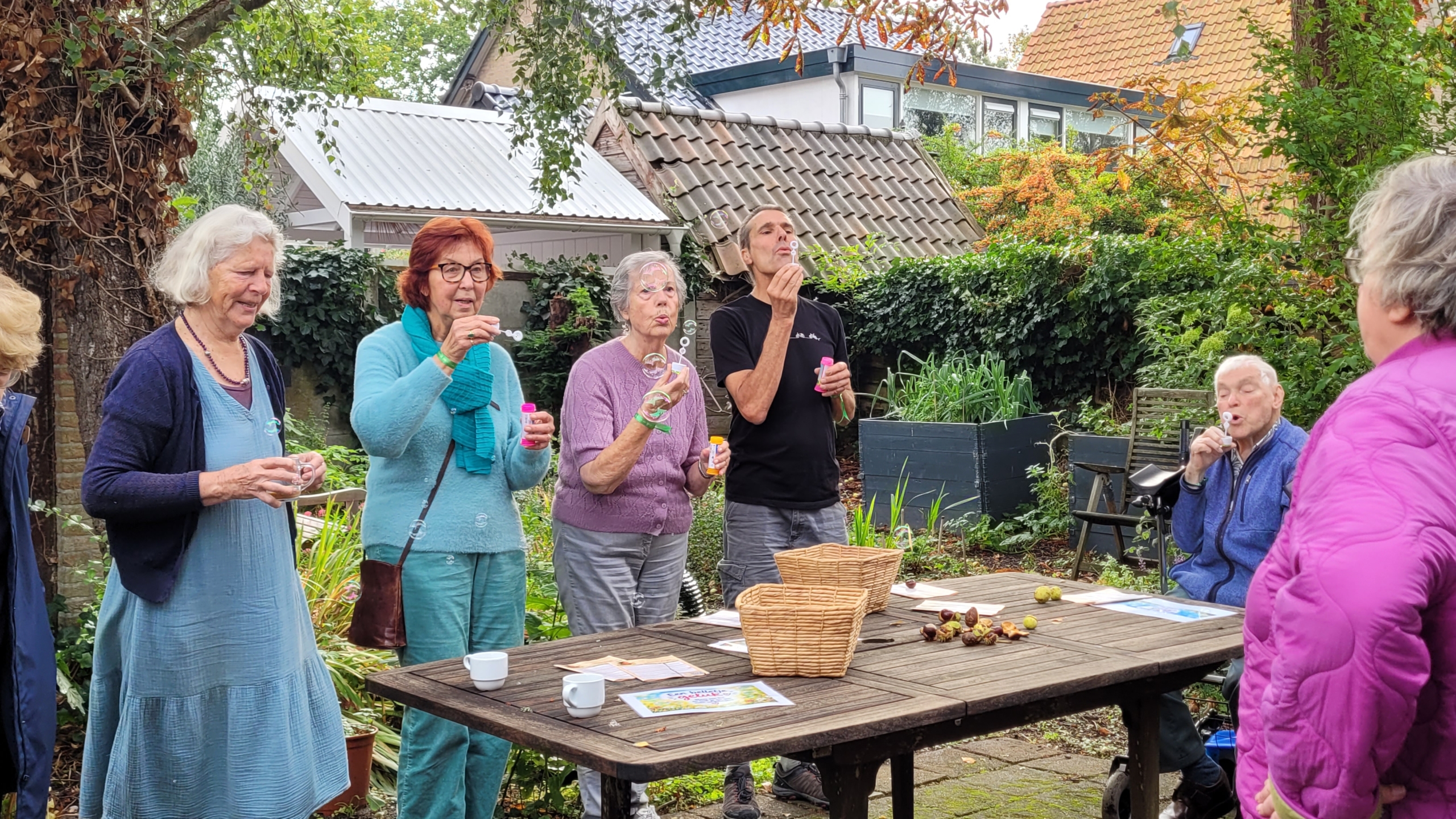 Groep mensen in een tuin blaast bellenblaas rond een houten tafel met manden en kopjes