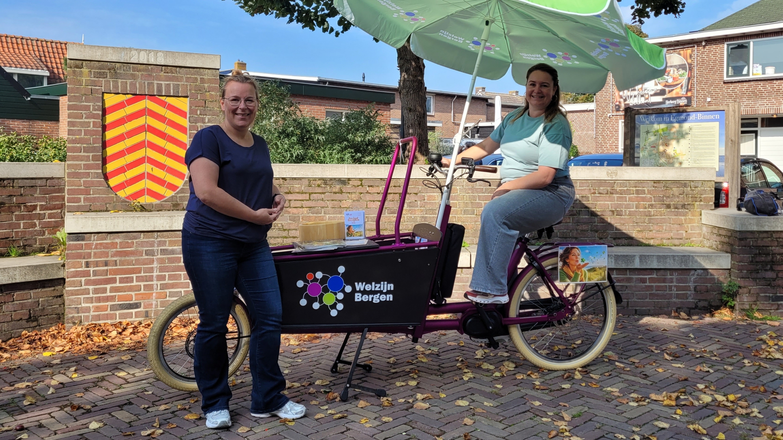 Twee vrouwen bij een paarse bakfiets met parasol en logo Welzijn Bergen, bij een muur met wapen en bord Egmond-Binnen