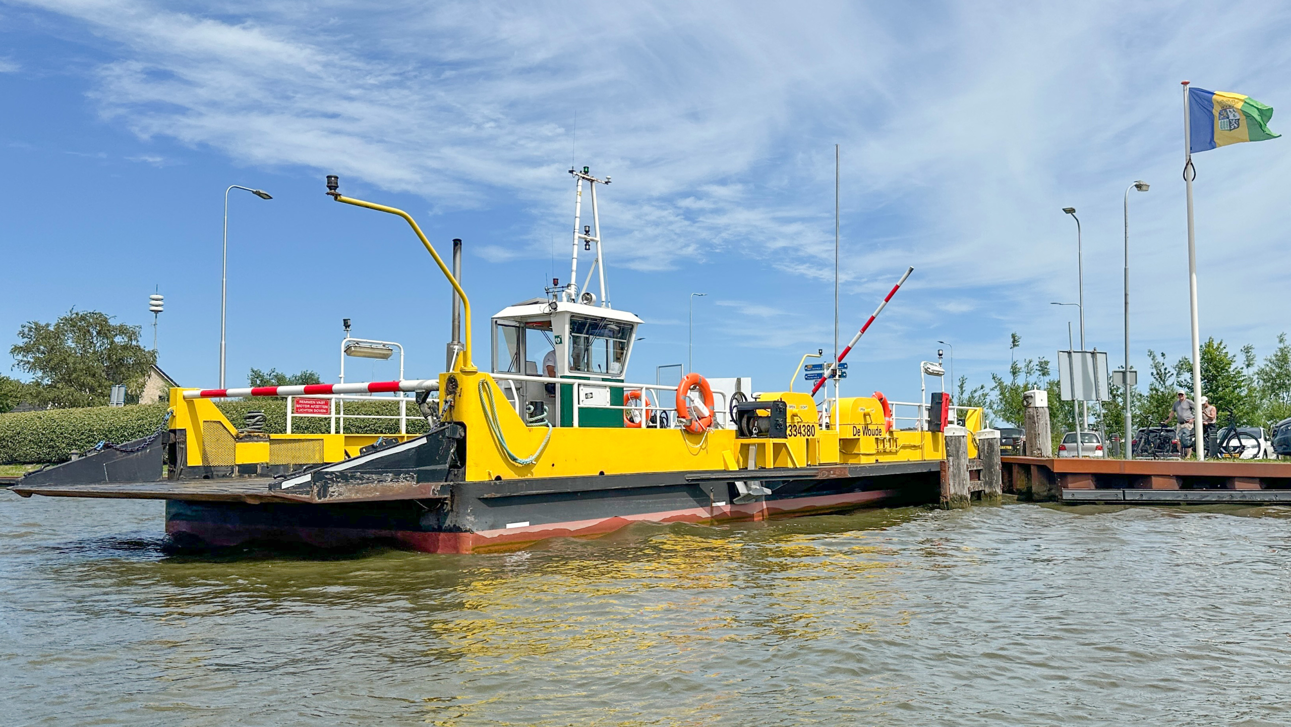 Gele veerpont “De Woude” aangemeerd aan de kade op het water, met slagbomen en een vlag op de achtergrond