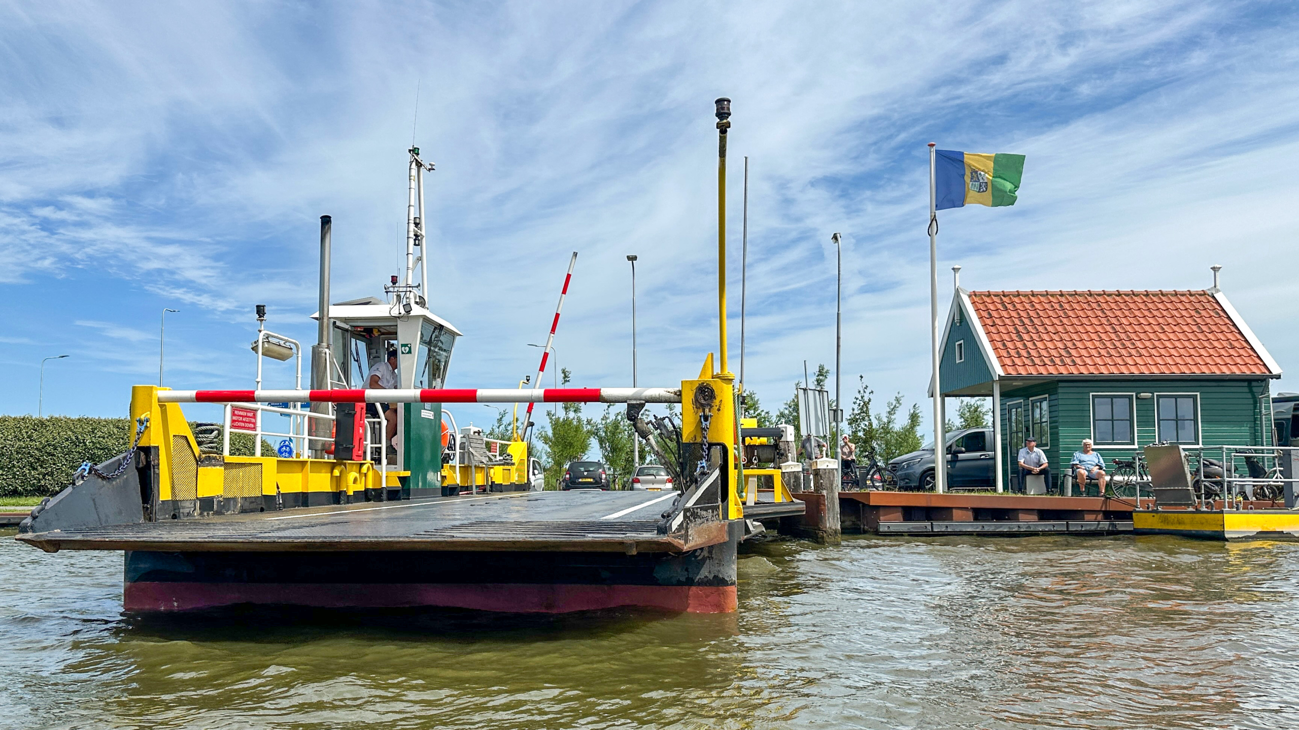 Pontveer met slagboom en oprit aan het water, met groen huisje en vlag op de kade onder blauwe lucht