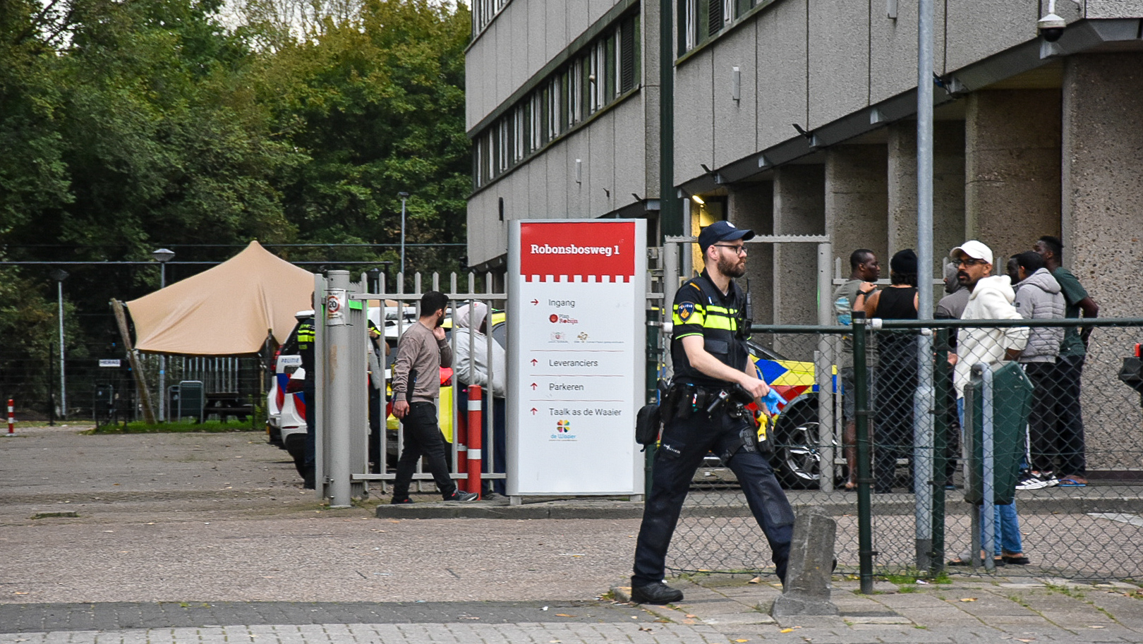 Politieagent loopt langs hek bij gebouw, met mensen in rij en bord Robonsbosweg 1 bij de ingang