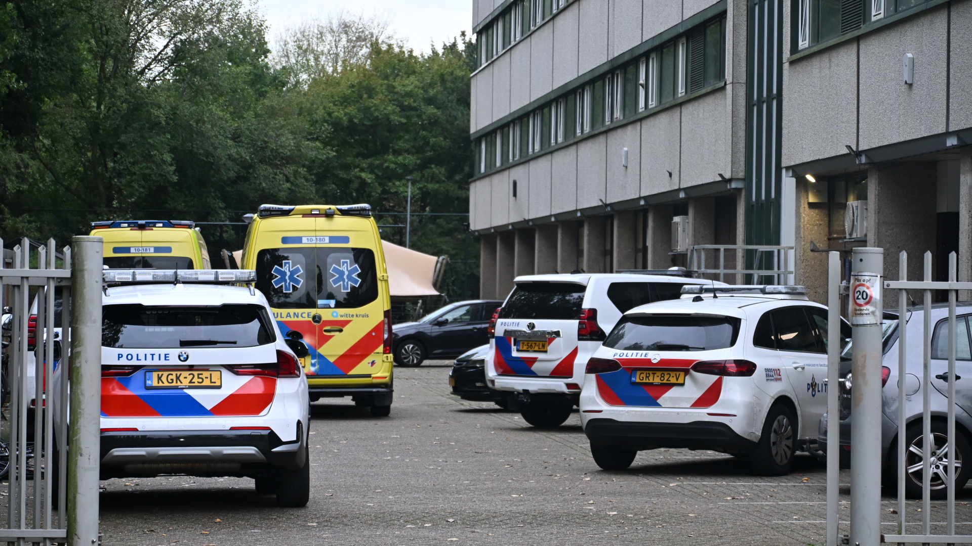 Twee politiewagens en twee ambulances geparkeerd bij een gebouw op een terrein met hekwerk