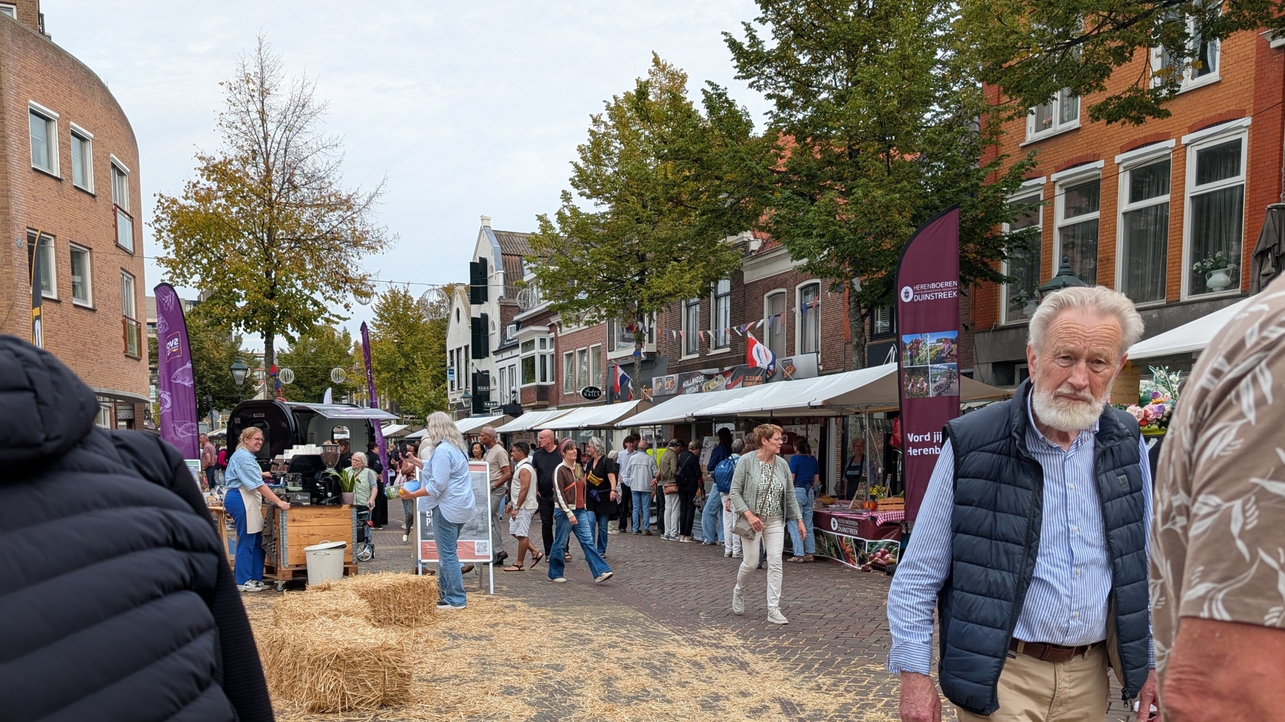 Drukke straatmarkt met kraampjes en spandoeken, strobalen op straat en bezoekers tussen gebouwen en bomen
