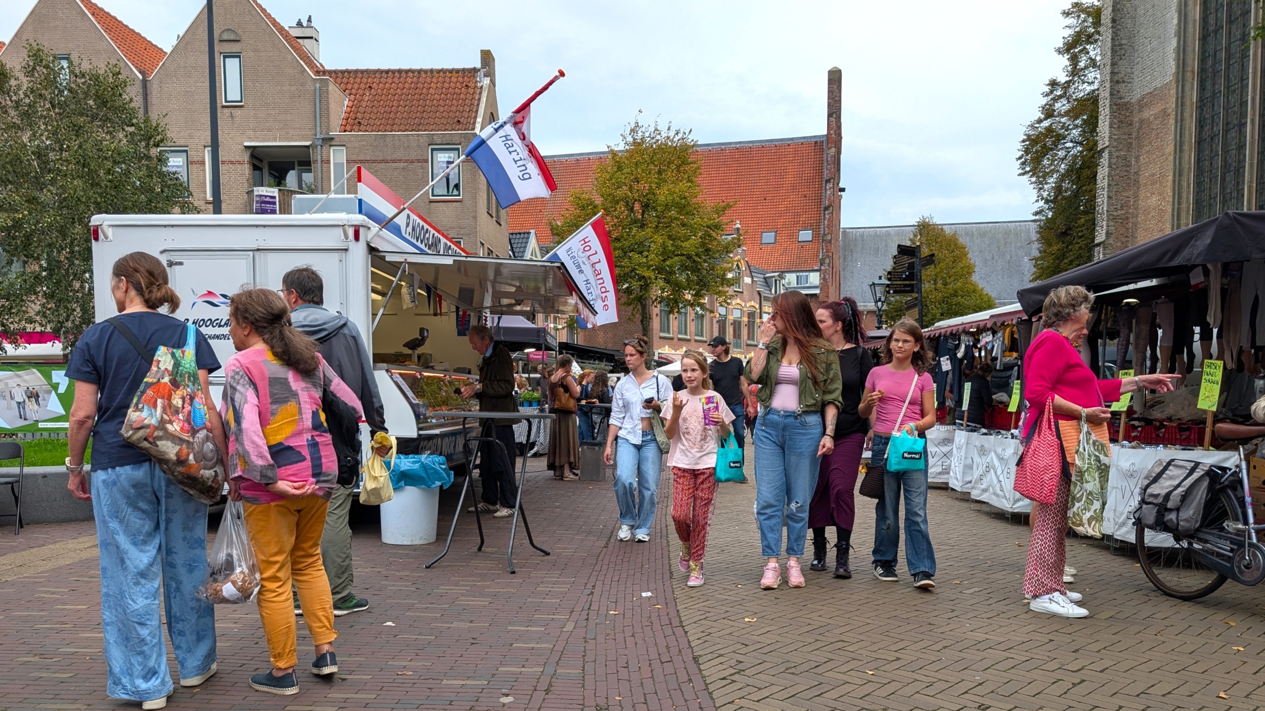 Drukke straatmarkt met kramen en winkelend publiek, met een viswagen en vlaggen met tekst Hoorn en Hollands