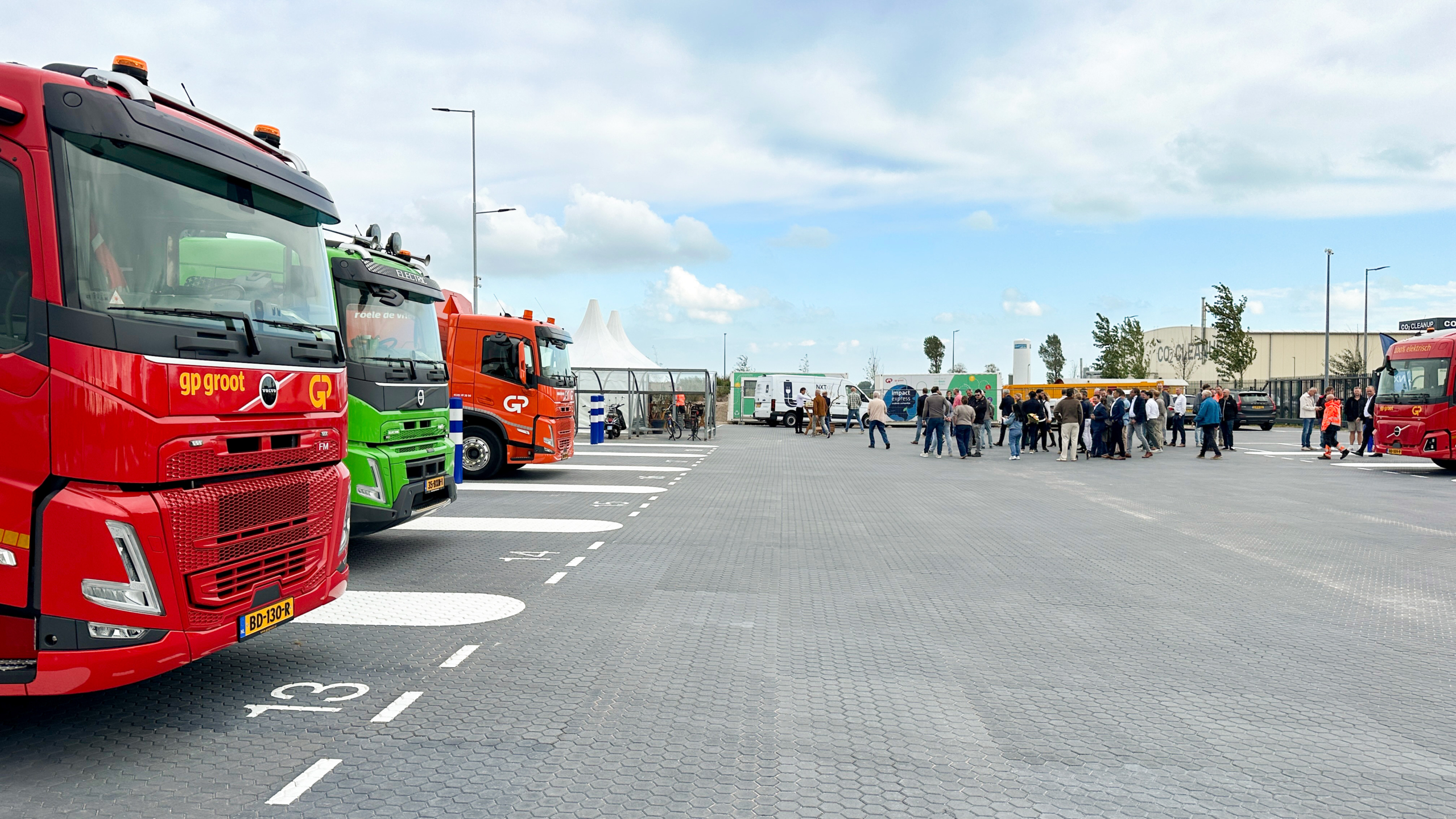 Rij gekleurde vrachtwagens op parkeerterrein, met groep mensen in de verte bij een evenemententent